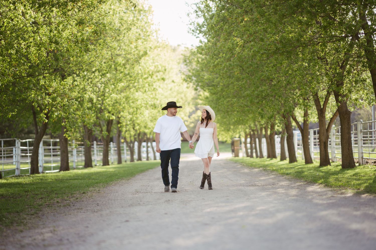 Couple in matching white outfits and cowboy hats stroll hand-in-hand along a scenic tree-lined country road.
