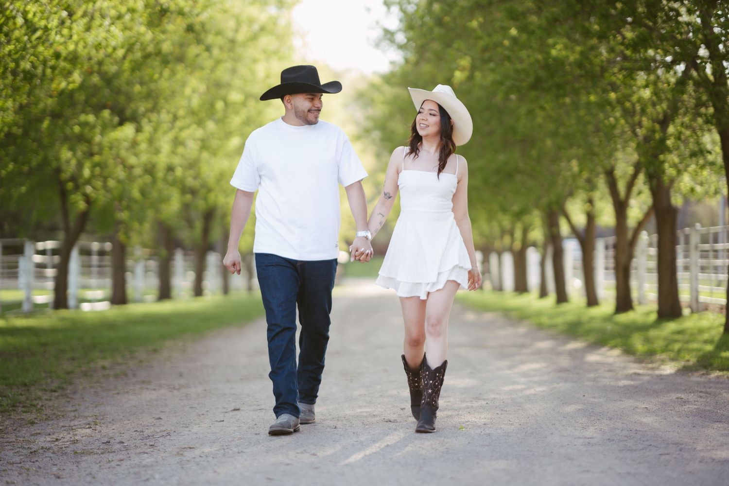 Couple in cowboy hats holding hands walking down a tree-lined path, wearing white outfits and cowboy boots.
