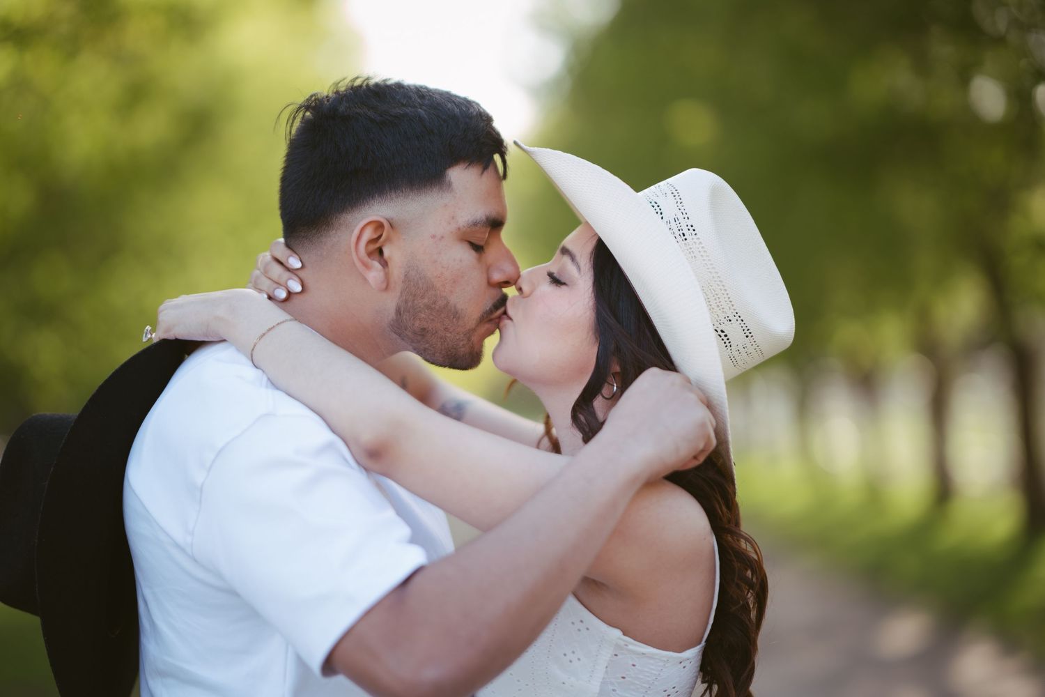 Couple sharing a romantic kiss outdoors, woman wearing white hat and dress, man in white shirt, green trees background.