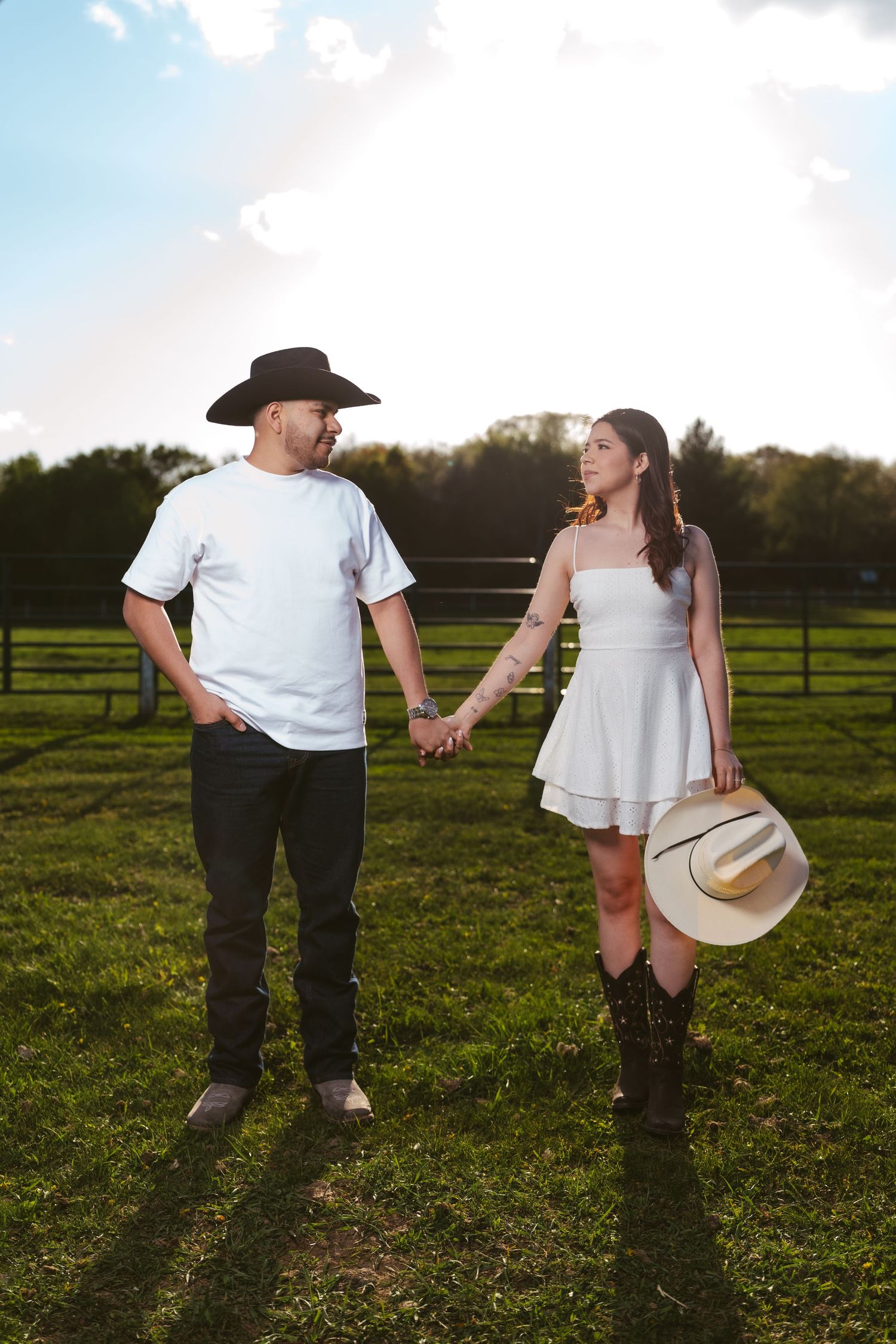 Couple holding hands in a sunny farm field, man in black cowboy hat and woman in white dress with boots.