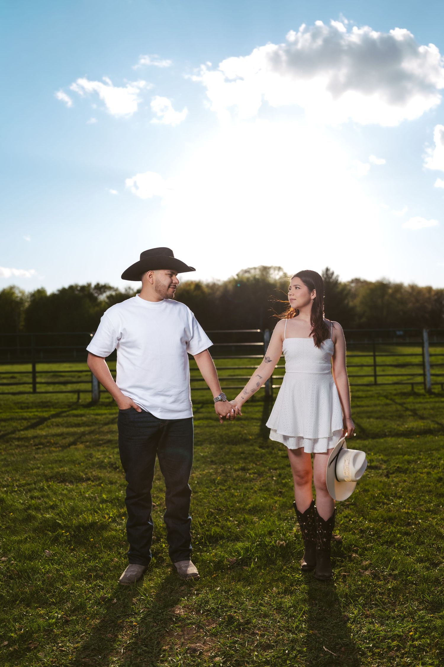 Couple holding hands in a sunny green pasture with fence and blue sky.