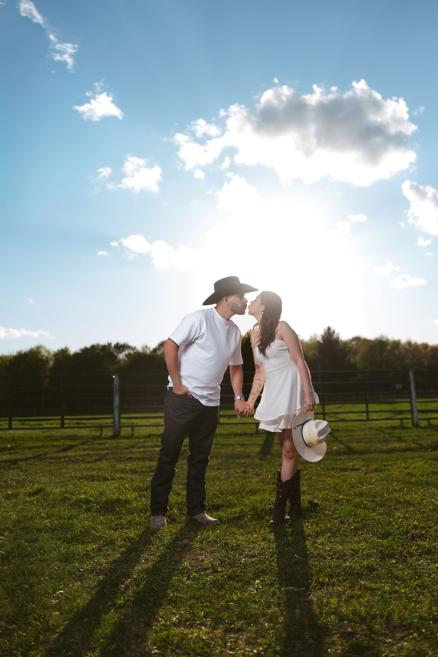 Couple kissing in sunlit green field, man in black cowboy hat, woman in white dress holding white hat.