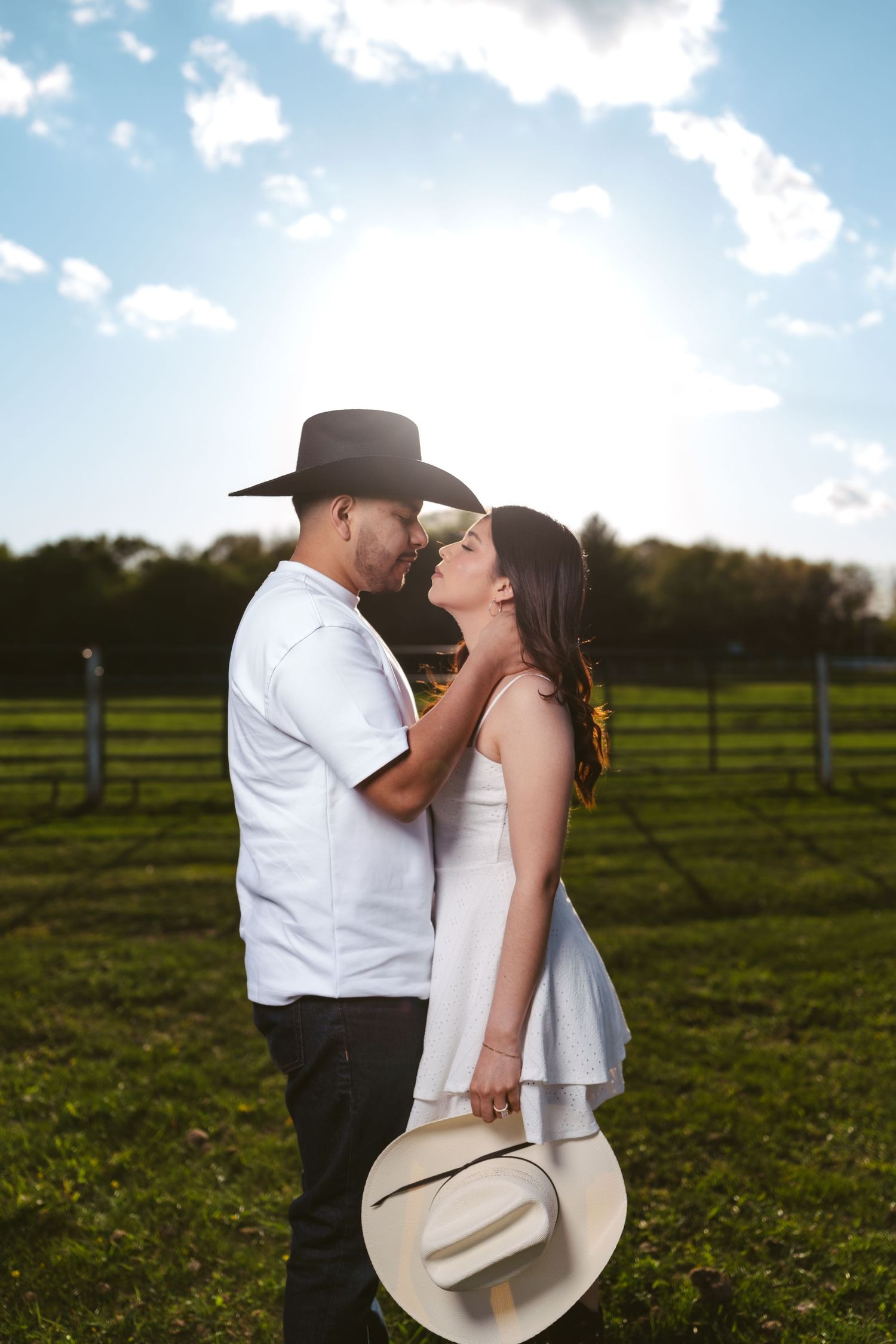 Couple sharing a kiss in a sunlit green field, man wearing black cowboy hat, woman holding white hat.