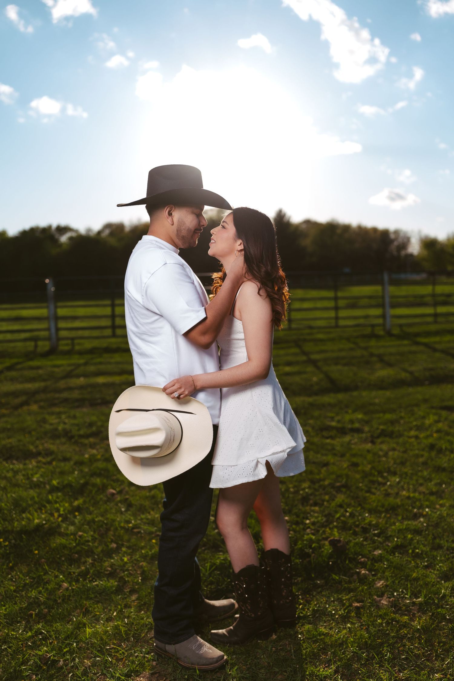 Couple embracing face-to-face in a sunny farm field, both wearing cowboy hats and white outfits.