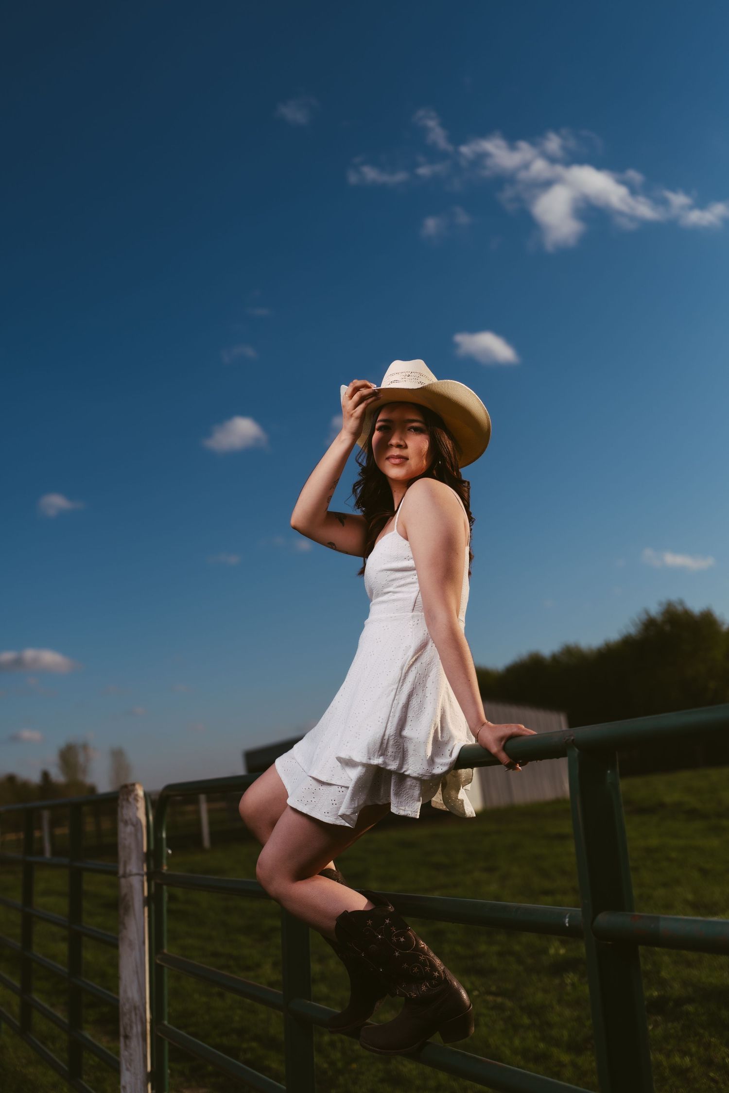 A woman in a white dress and cowboy hat sits on a farm fence under a vivid blue sky.