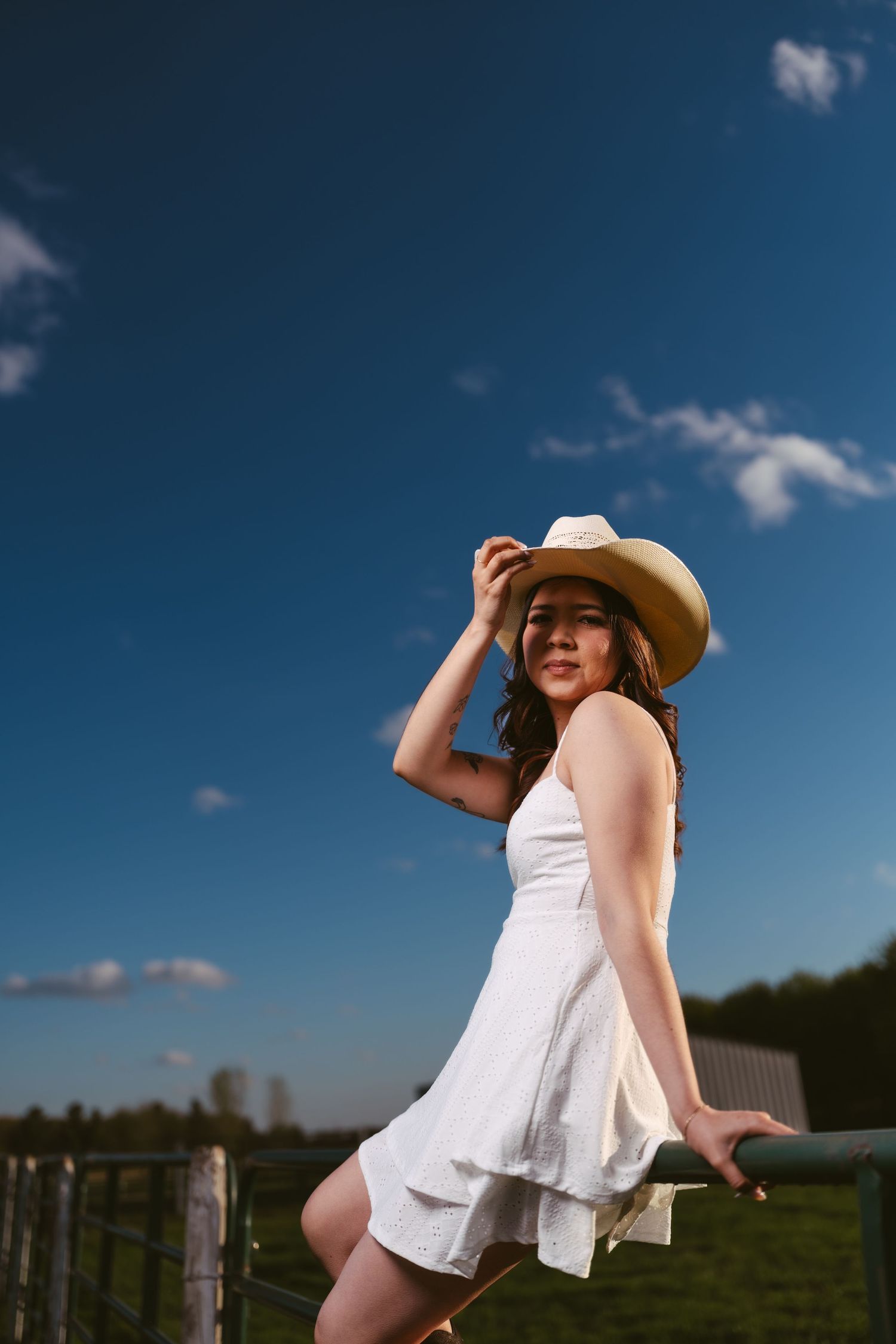 Young woman in white dress and cowboy hat posing by a fence under a vivid blue sky on a sunny day.