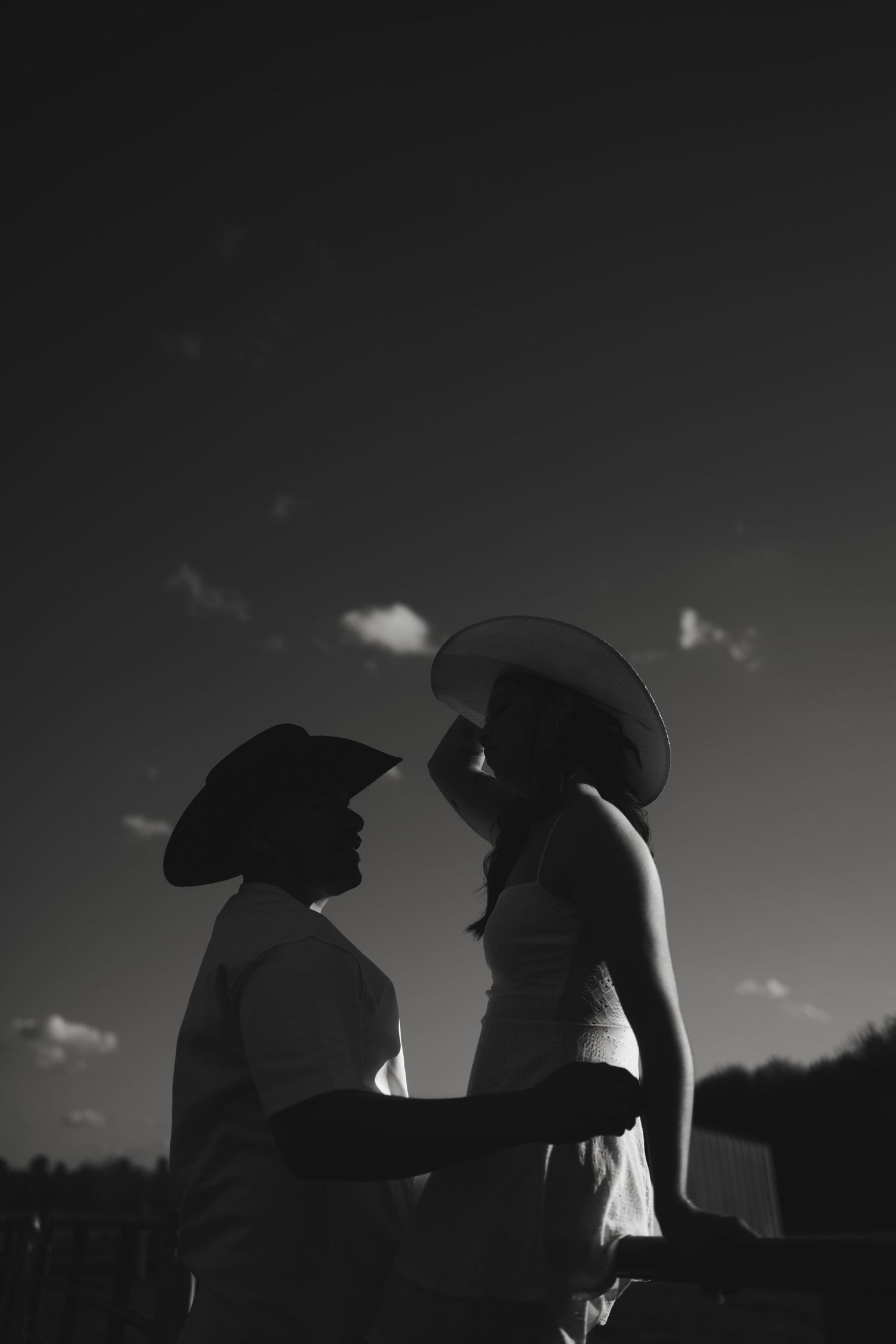 Black and white silhouette of romantic couple in cowboy hats facing each other against dramatic sky.