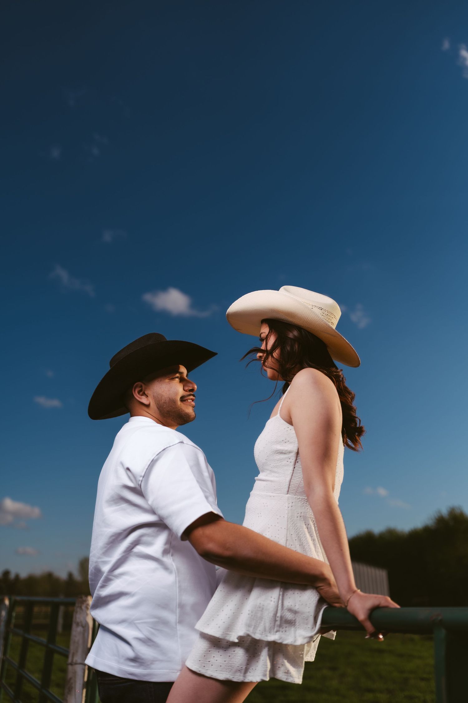 Smiling couple in cowboy hats holding hands by a farm fence under a deep blue sky.