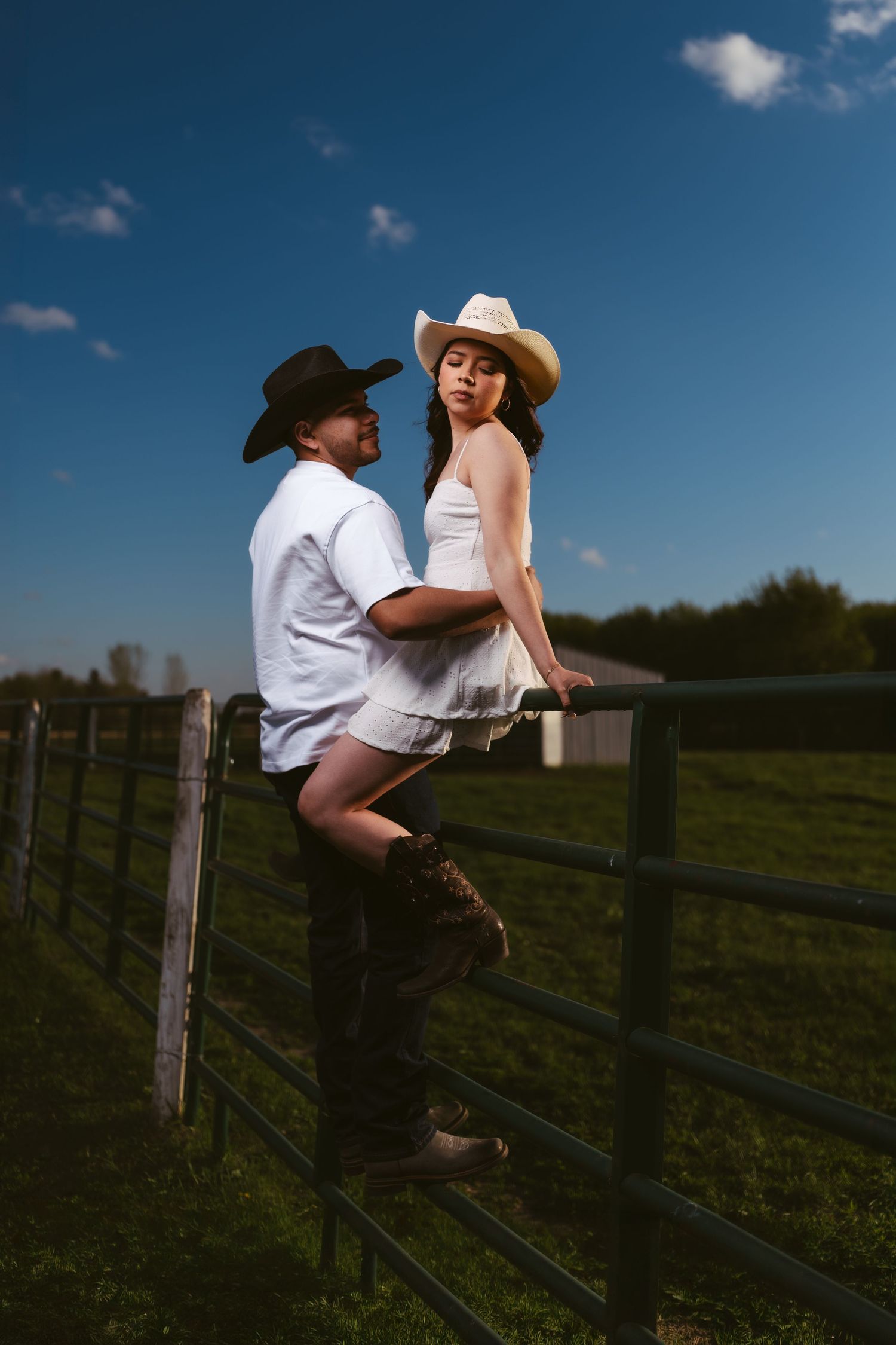Couple in cowboy hats pose on a farm fence under a deep blue sky.