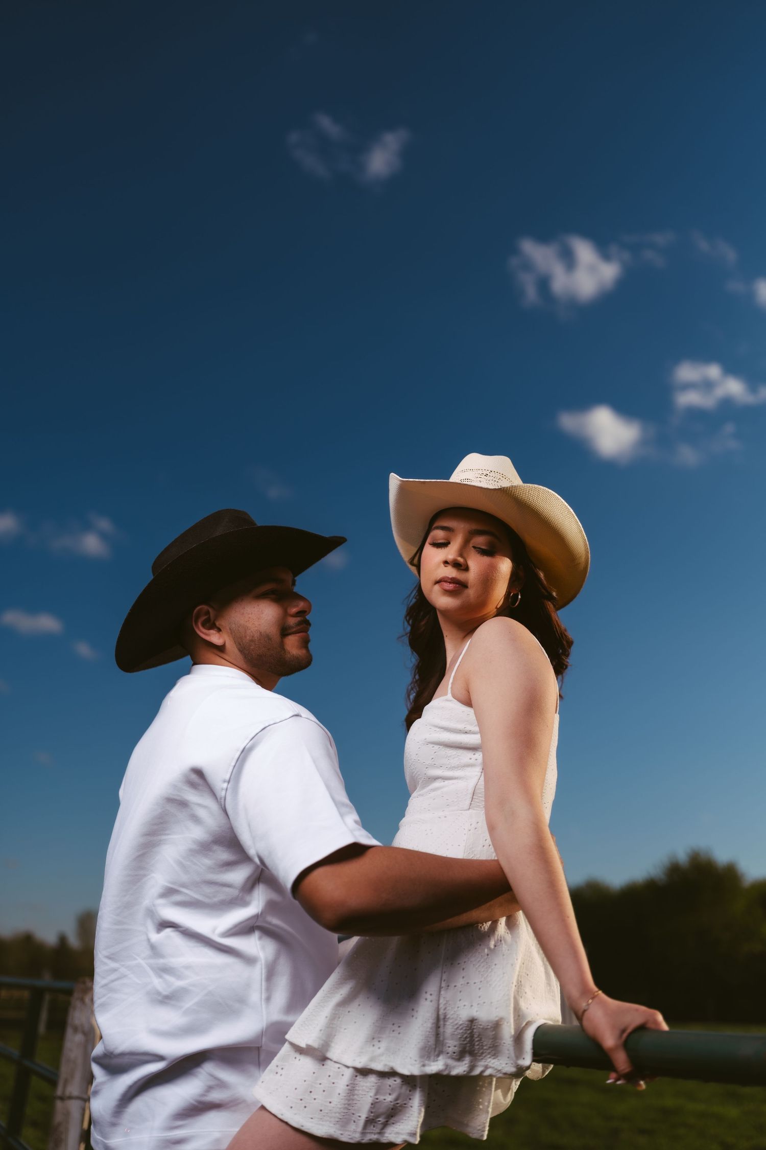 Couple embracing near a farm fence under a vivid blue sky, both wearing cowboy hats in white outfits.