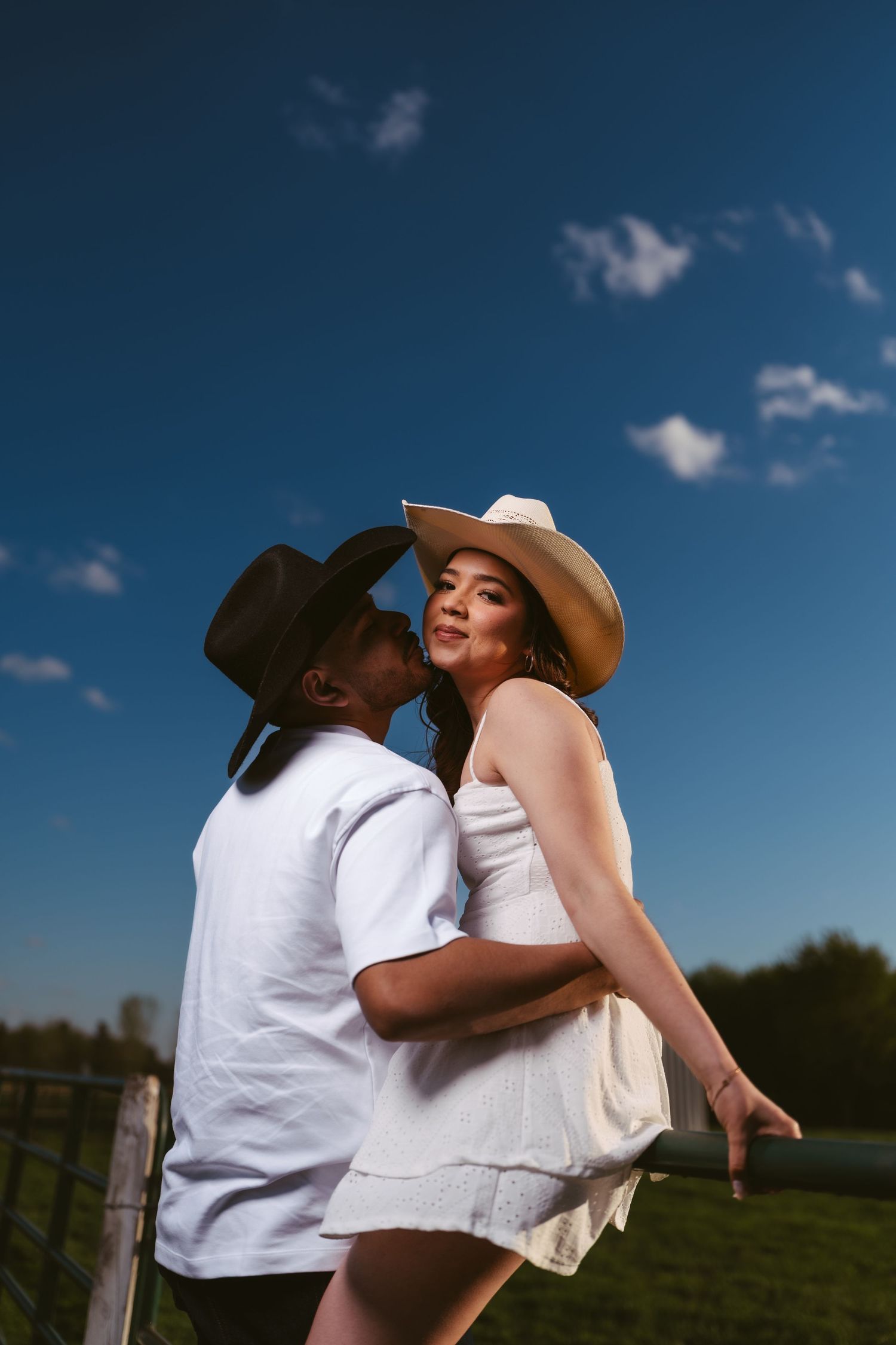 A couple in cowboy hats embraces near a farm fence with a bright blue sky backdrop.