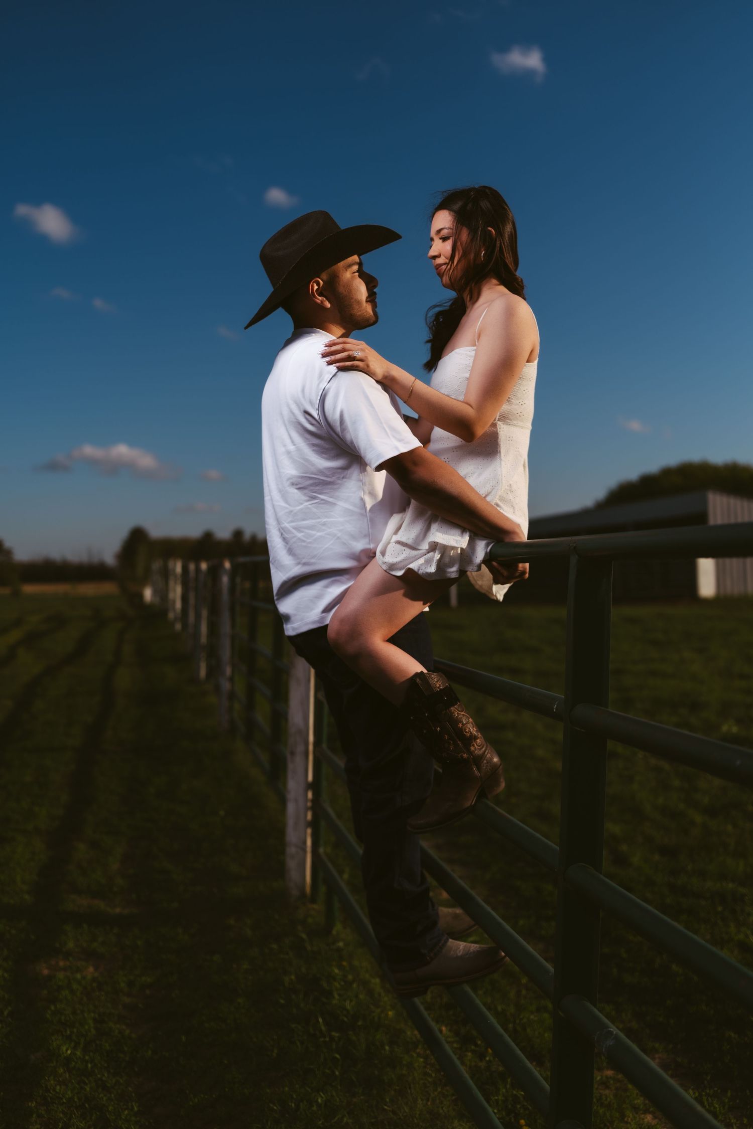 A romantic couple gazes at each other on a farm fence at golden hour on a ranch.