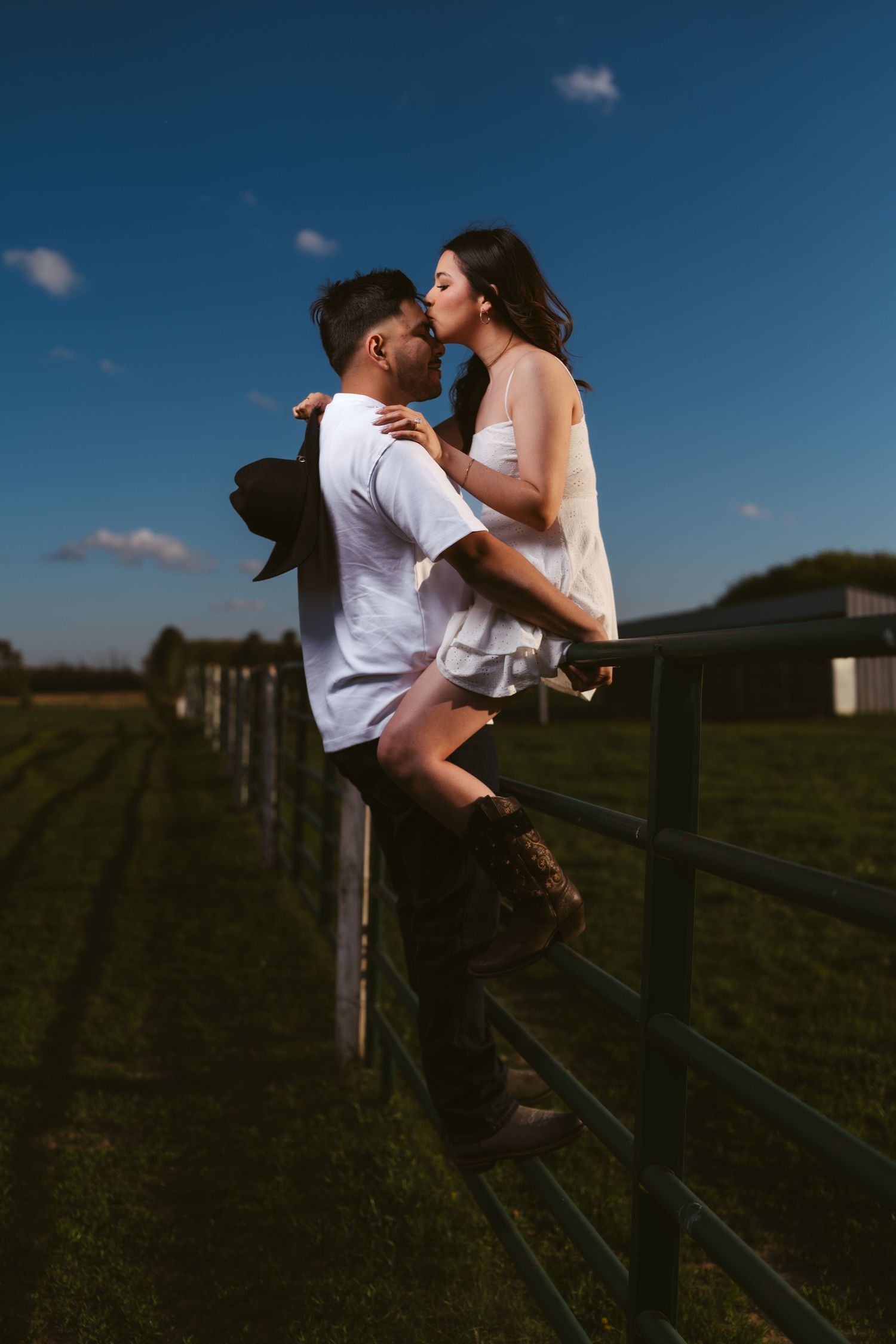 Romantic couple kissing on a farm fence at sunset, woman in white dress and cowboy boots sitting on rail.