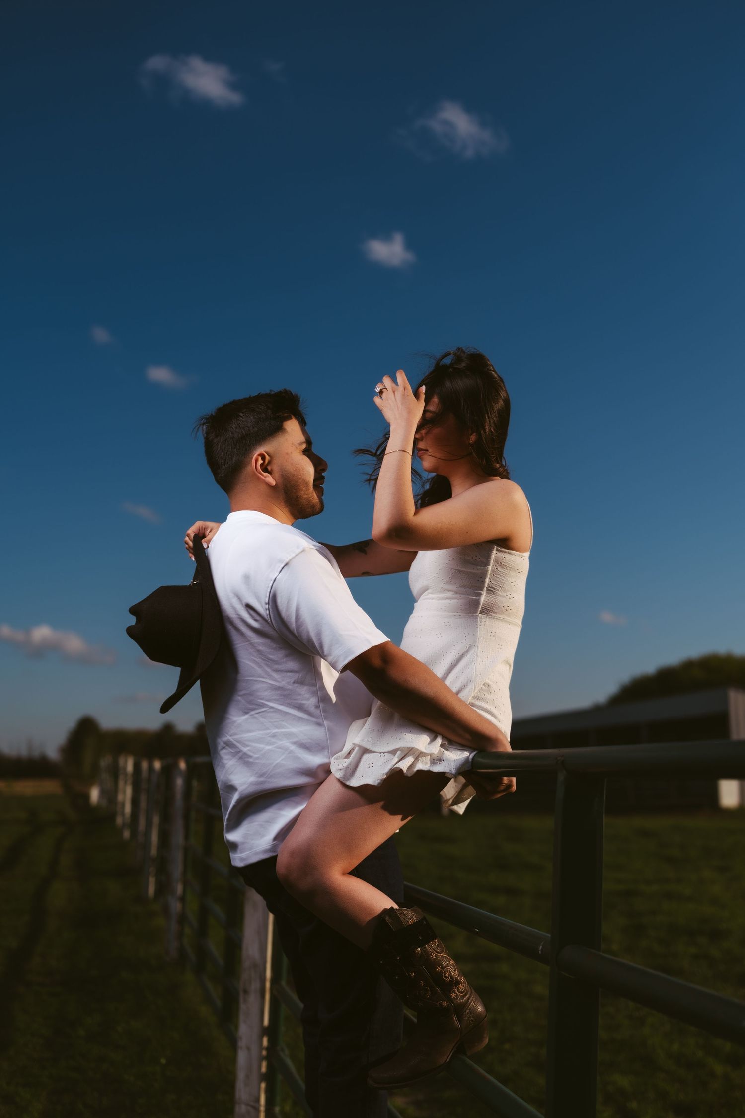 Woman sitting on farm fence embracing man in white shirt against dramatic blue sky at golden hour.