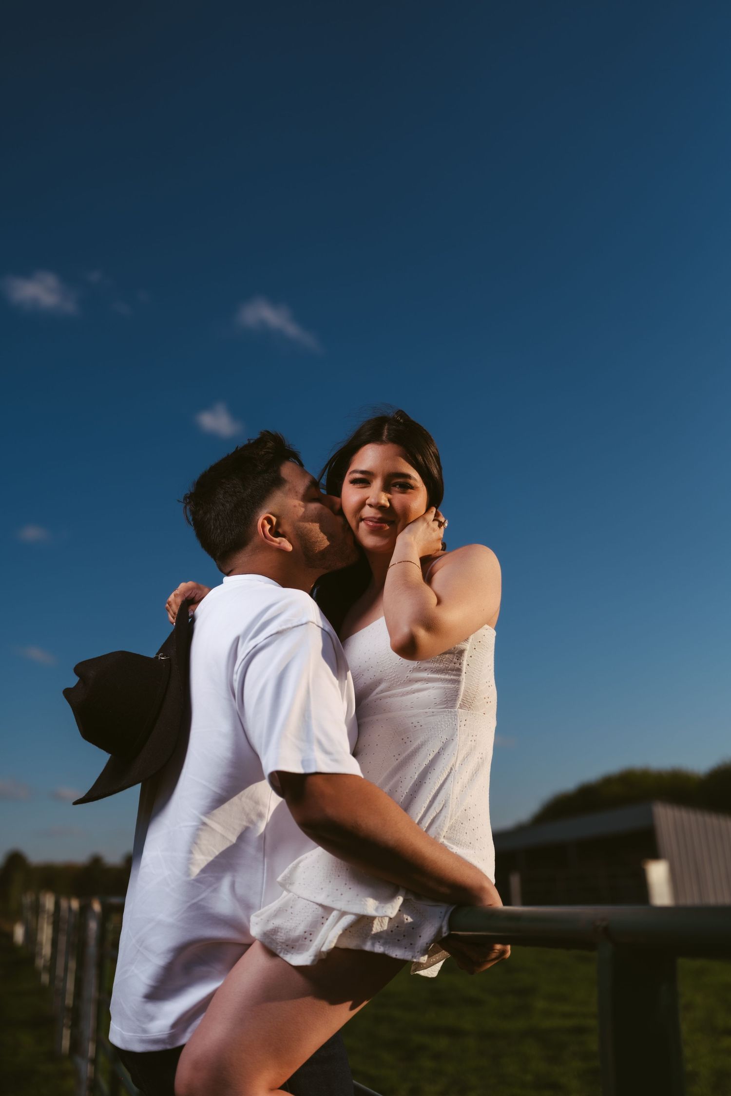 Romantic couple embracing at sunset by a farm fence under a vivid blue sky.