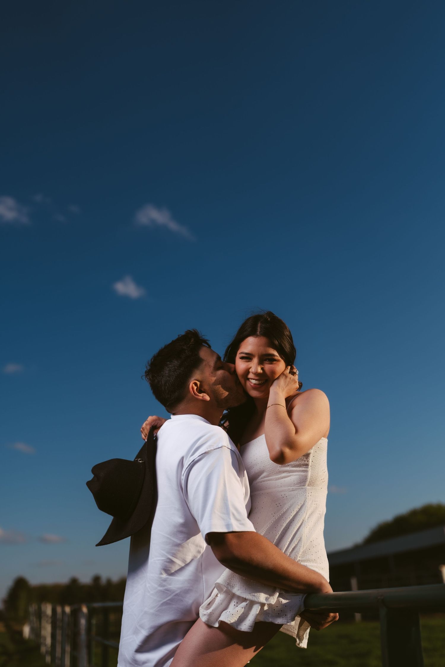 Man kissing smiling woman on cheek while holding her at a farm fence during sunset.