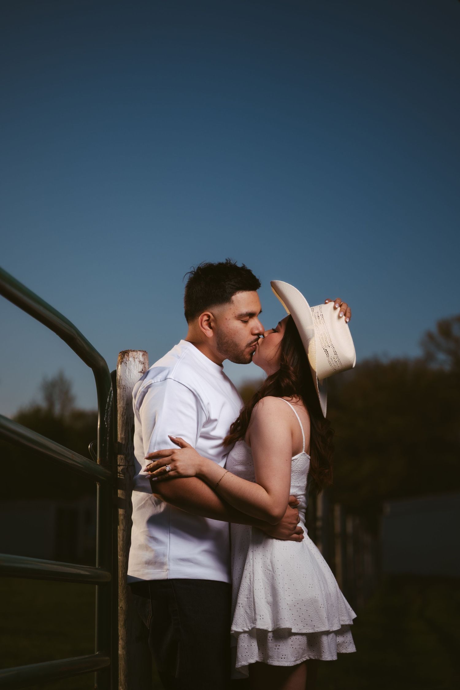 Couple sharing a passionate kiss by a farm gate at dusk, woman in white dress and cowboy hat embracing man.