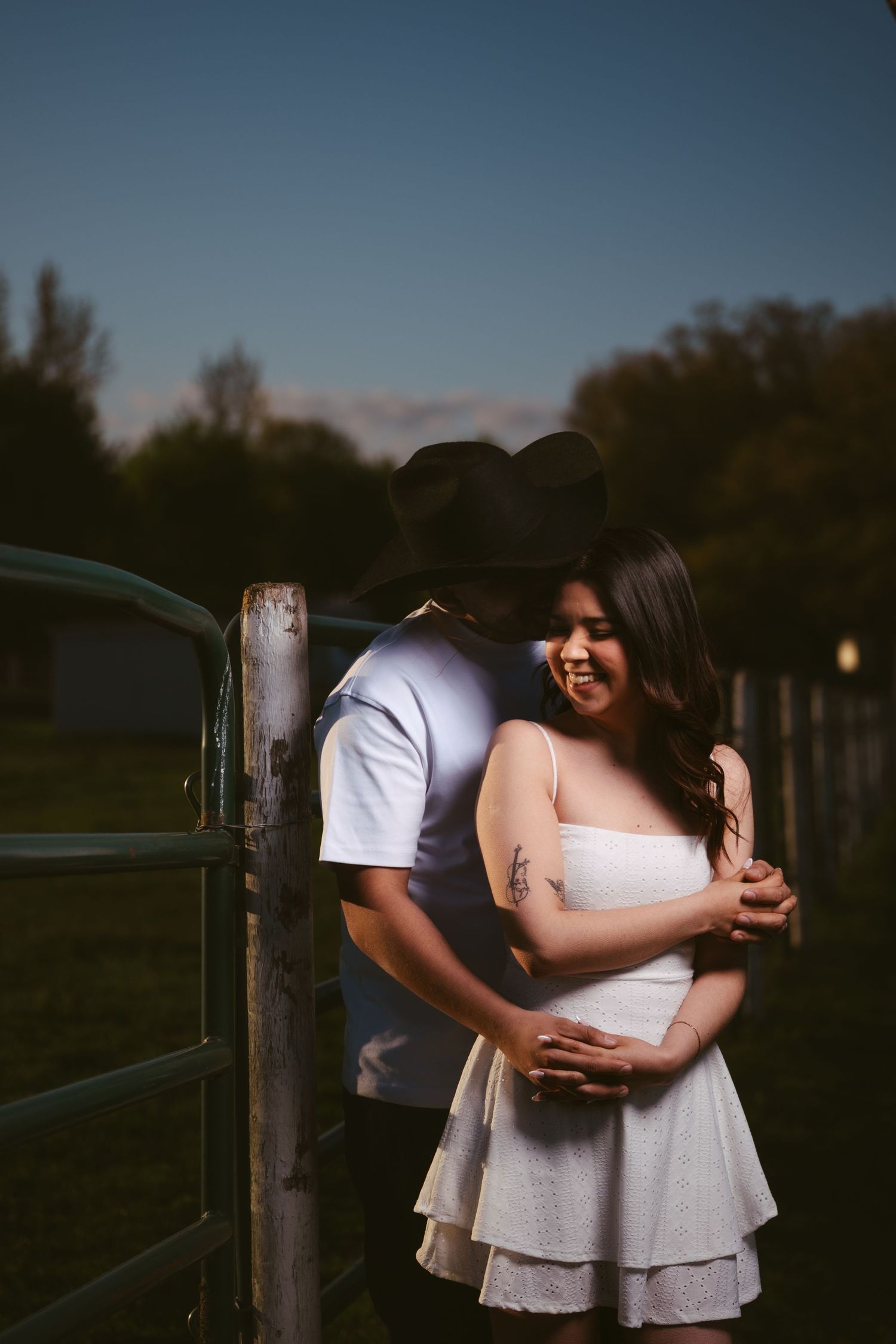 Couple embracing by a rustic farm gate at dusk, man in black cowboy hat nuzzling woman.