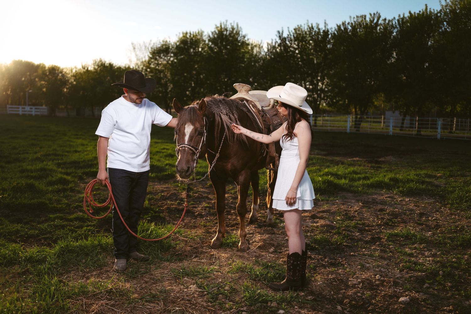 Couple standing with a brown horse in a sunny field, woman in white dress and cowboy hat stroking the horse.