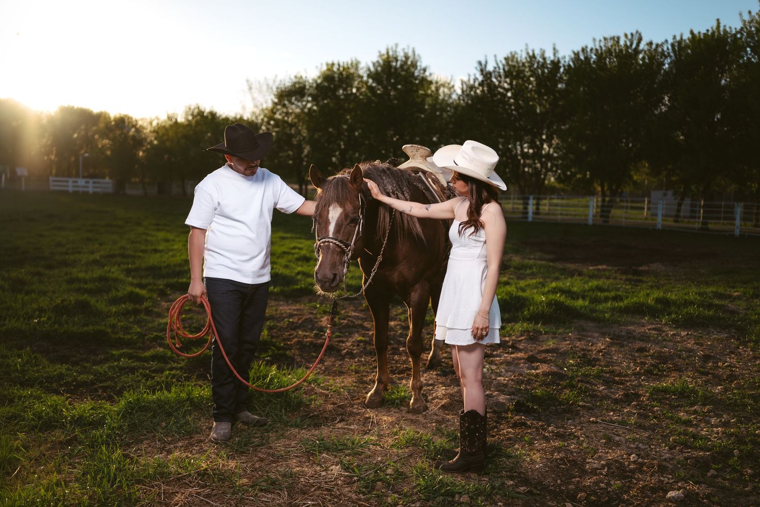 Couple in white outfits petting a brown horse at golden hour in a sunny green pasture.