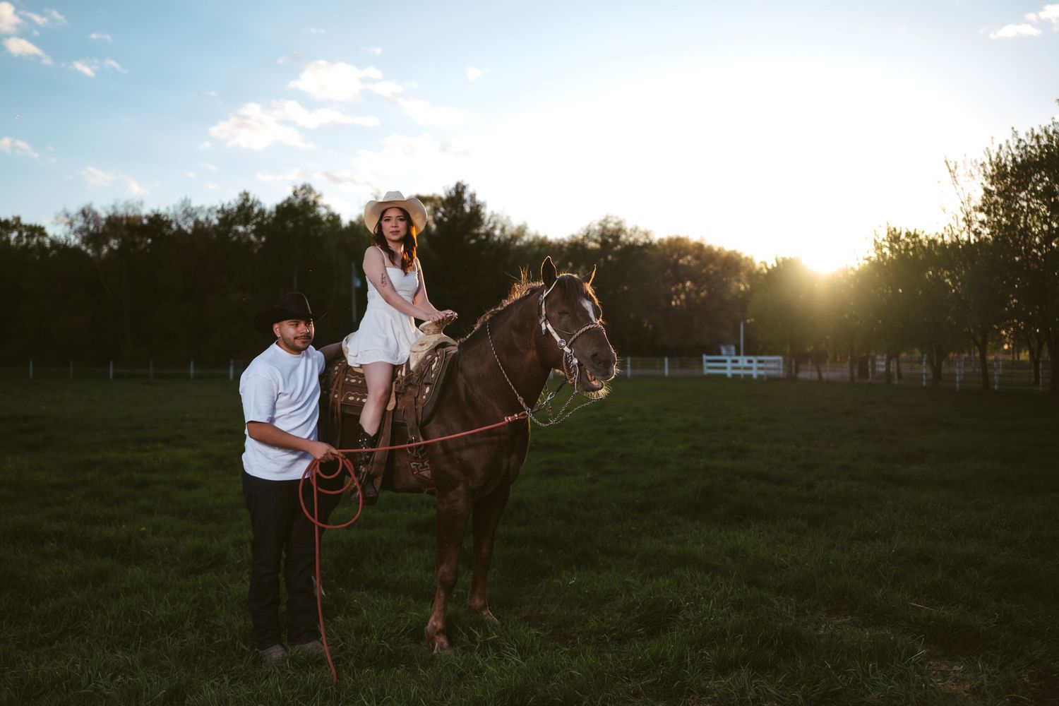 A woman in a white dress and cowboy hat rides a horse at sunset on a green farm.
