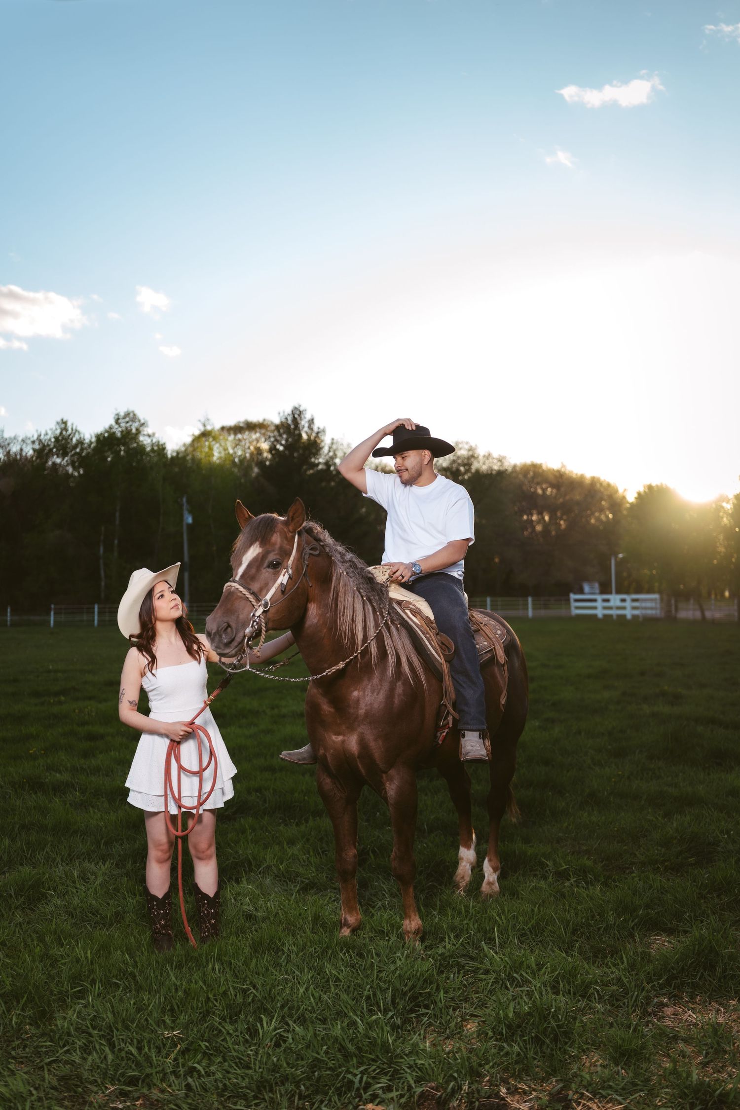 Woman in white dress holding reins as man in cowboy hat sits on brown horse at golden sunset.