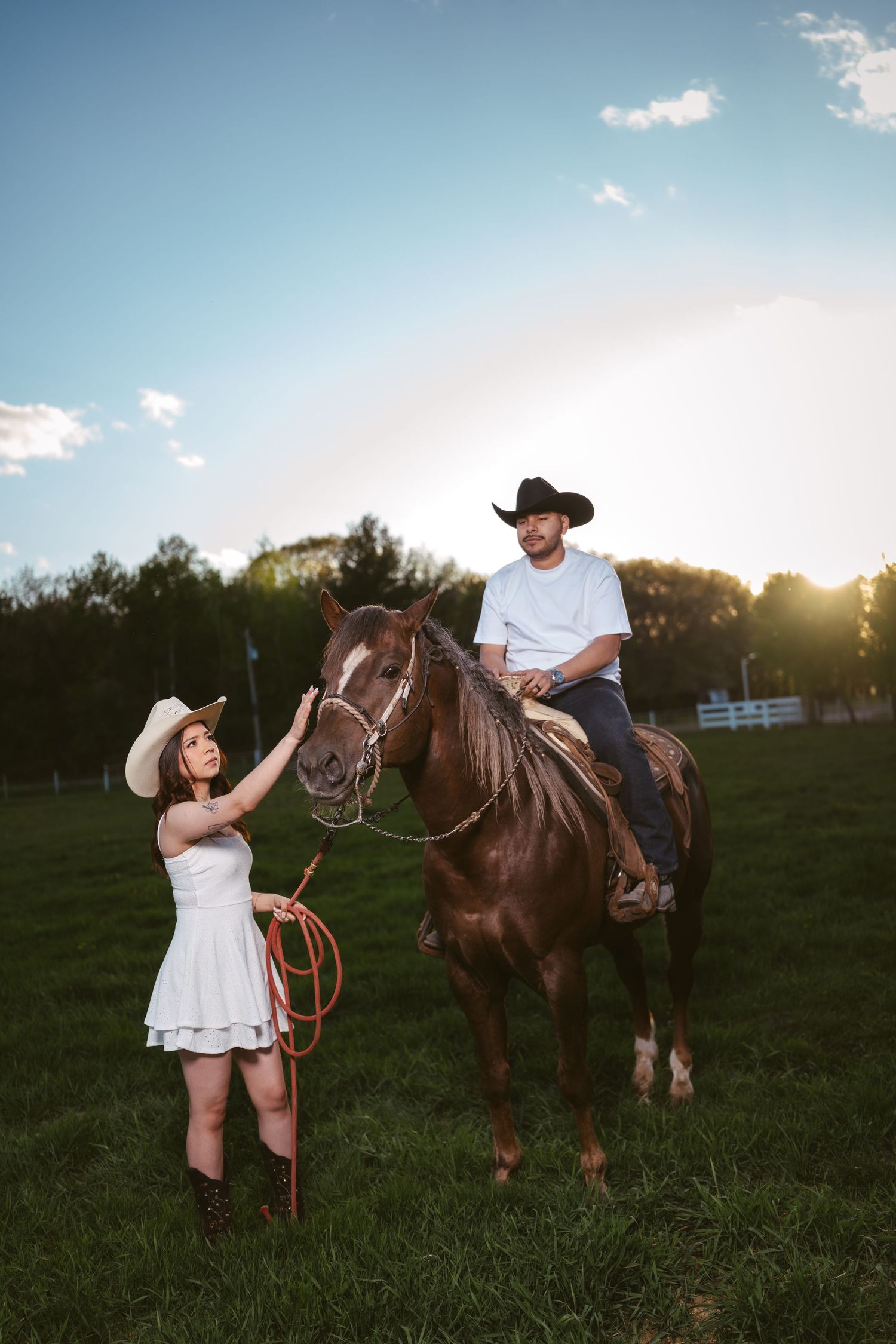 A cowboy on horseback looks down at a woman in white dress and cowboy hat holding a lasso at sunset.