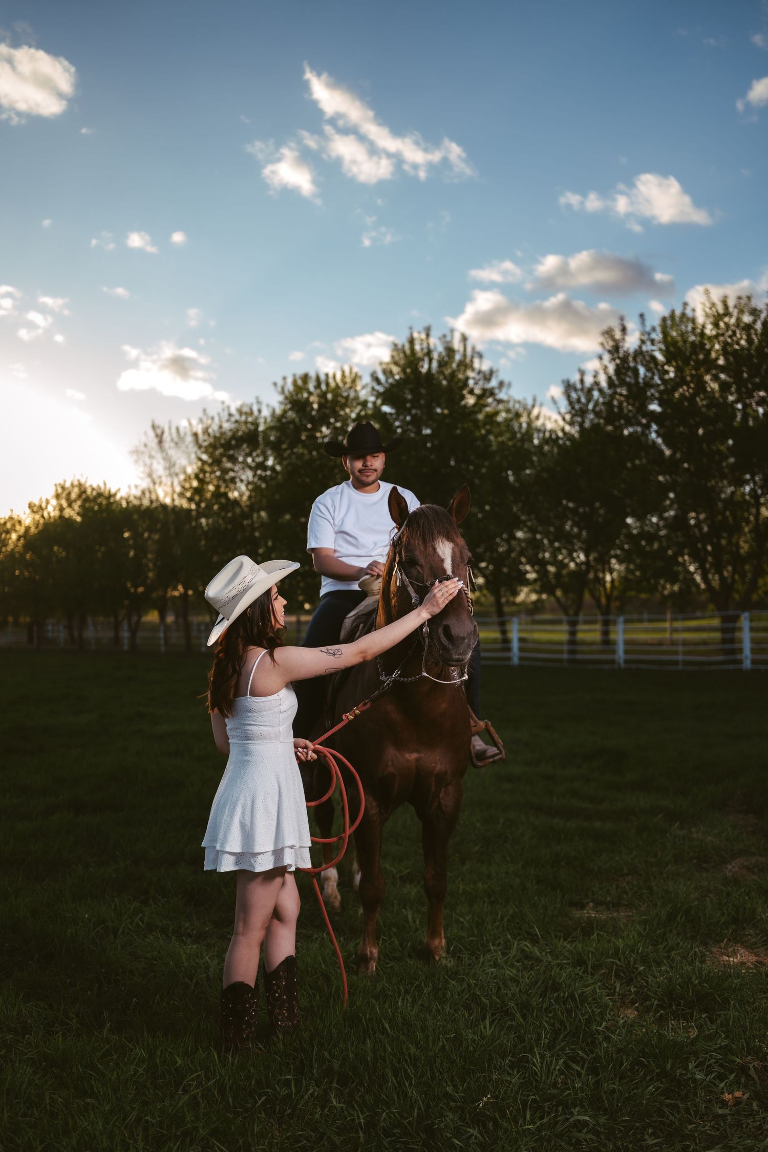 A woman in a white hat pets a horse as a man sits atop it in a sunny green field.