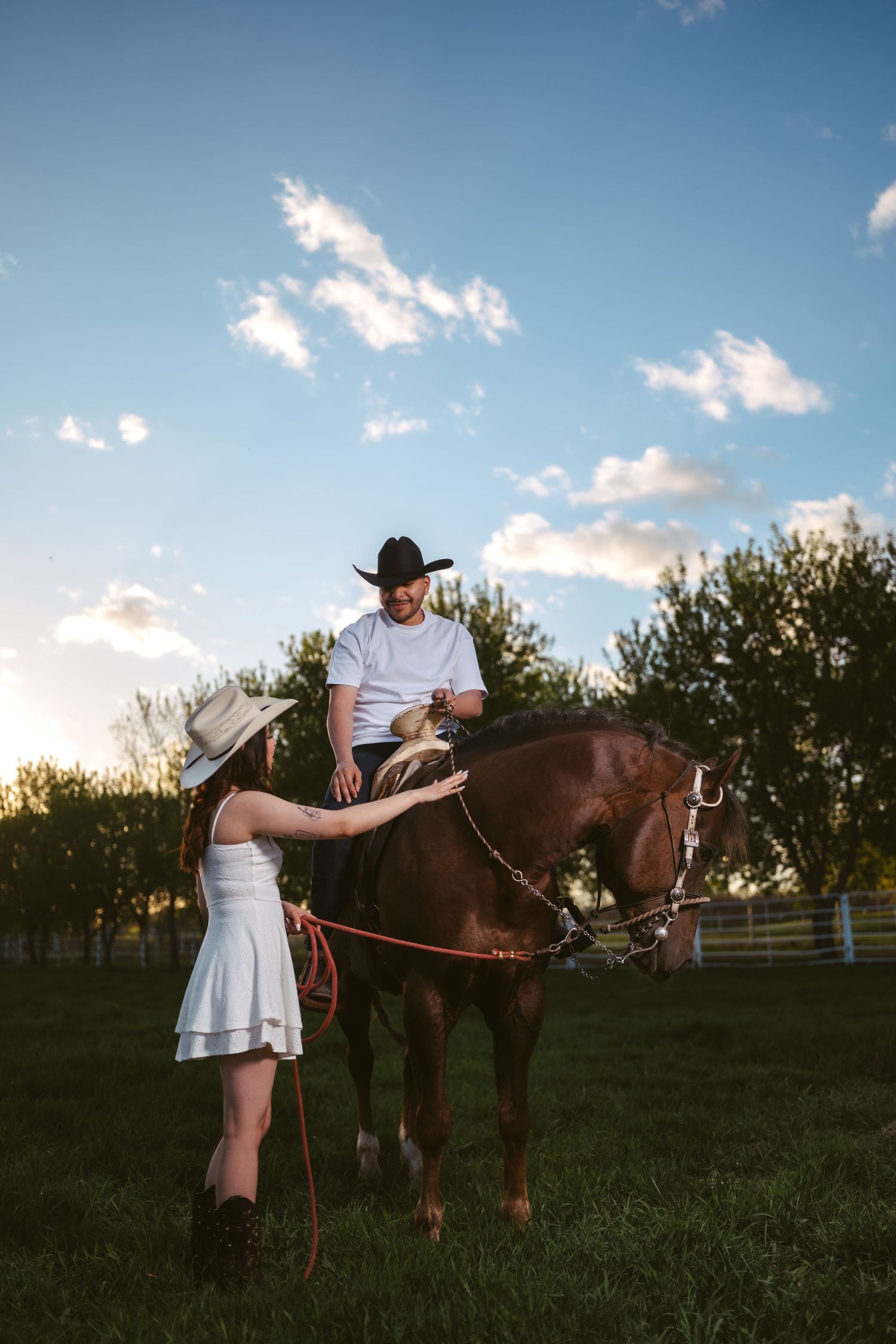 A woman in white dress and cowboy hat leads a horse with a man riding it across a green field at dusk.