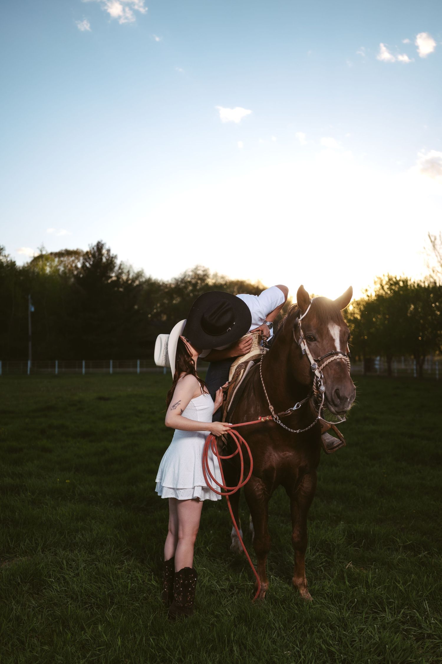 Couple in cowboy hats kissing beside a brown horse at golden sunset.