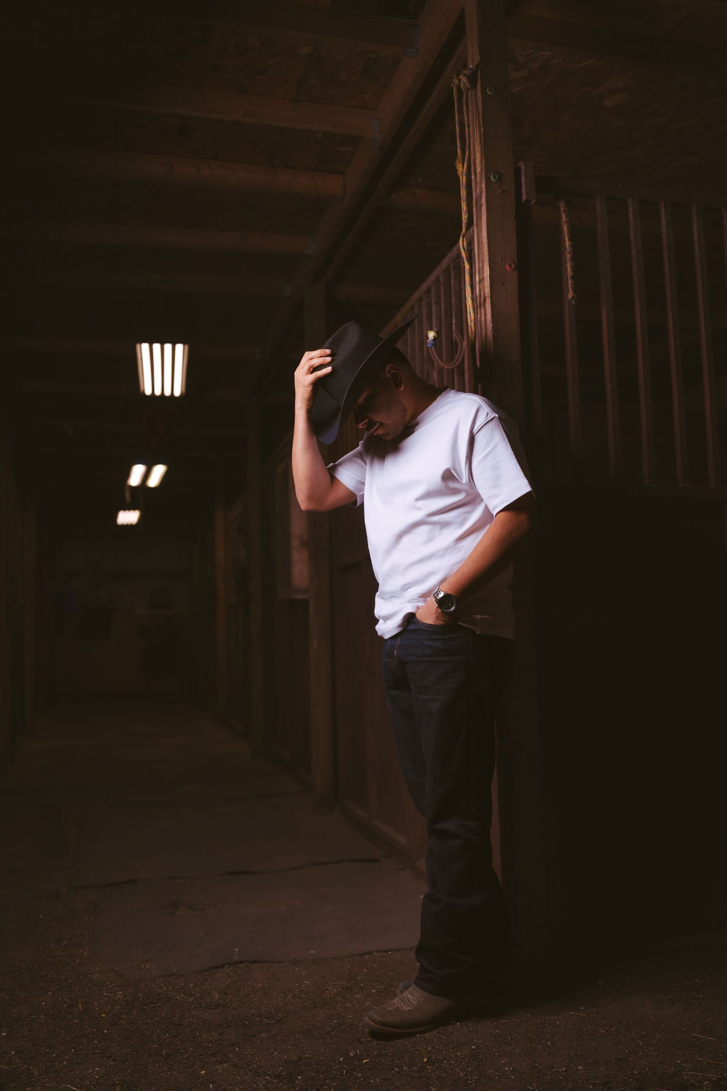 A man in a white shirt and black cowboy hat leans against a dark rustic barn wall.