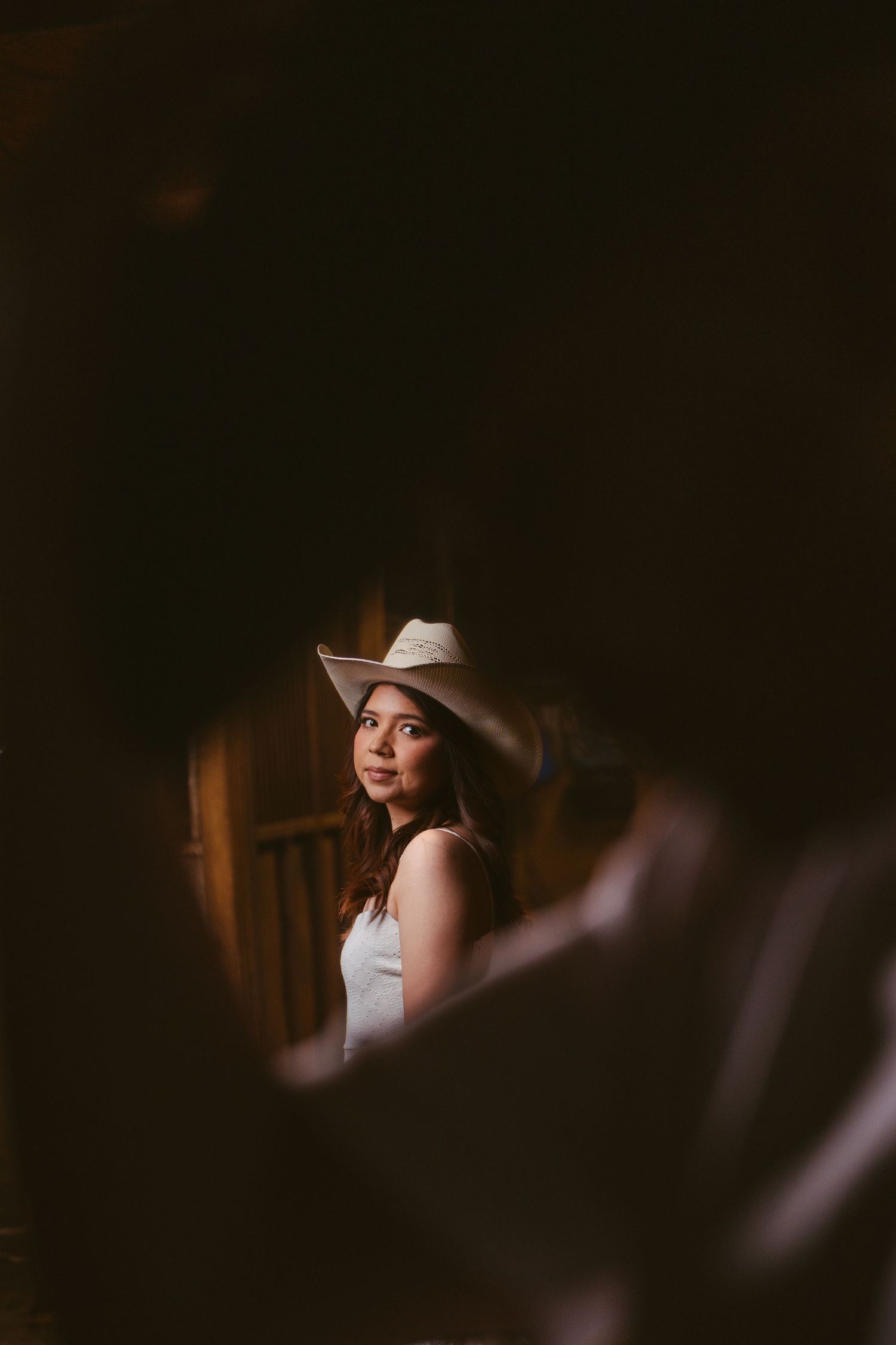A beautiful young woman in a white cowboy hat gazes softly in a dimly lit rustic barn setting.