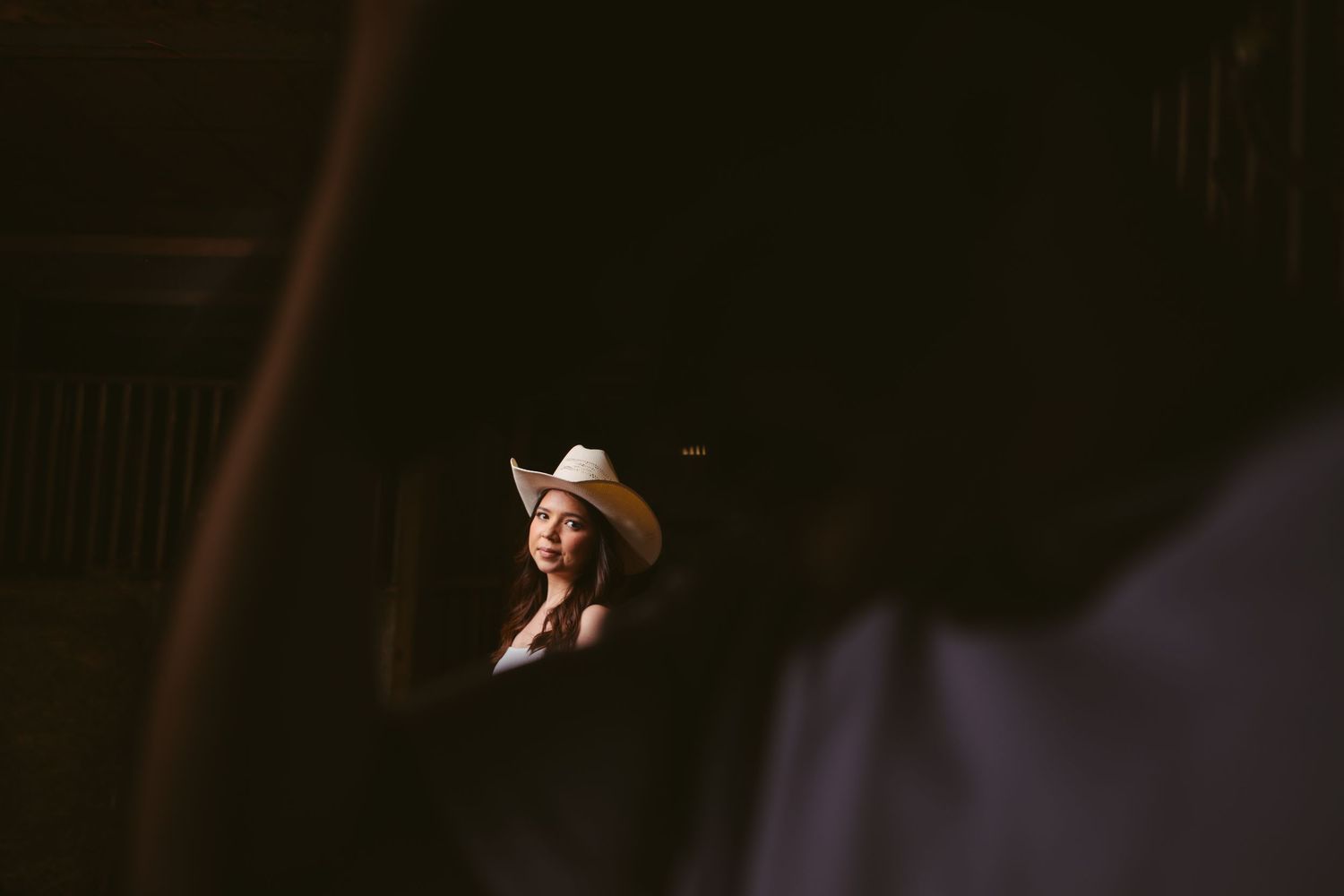 Woman in white cowboy hat gazes softly in a dimly lit barn setting.