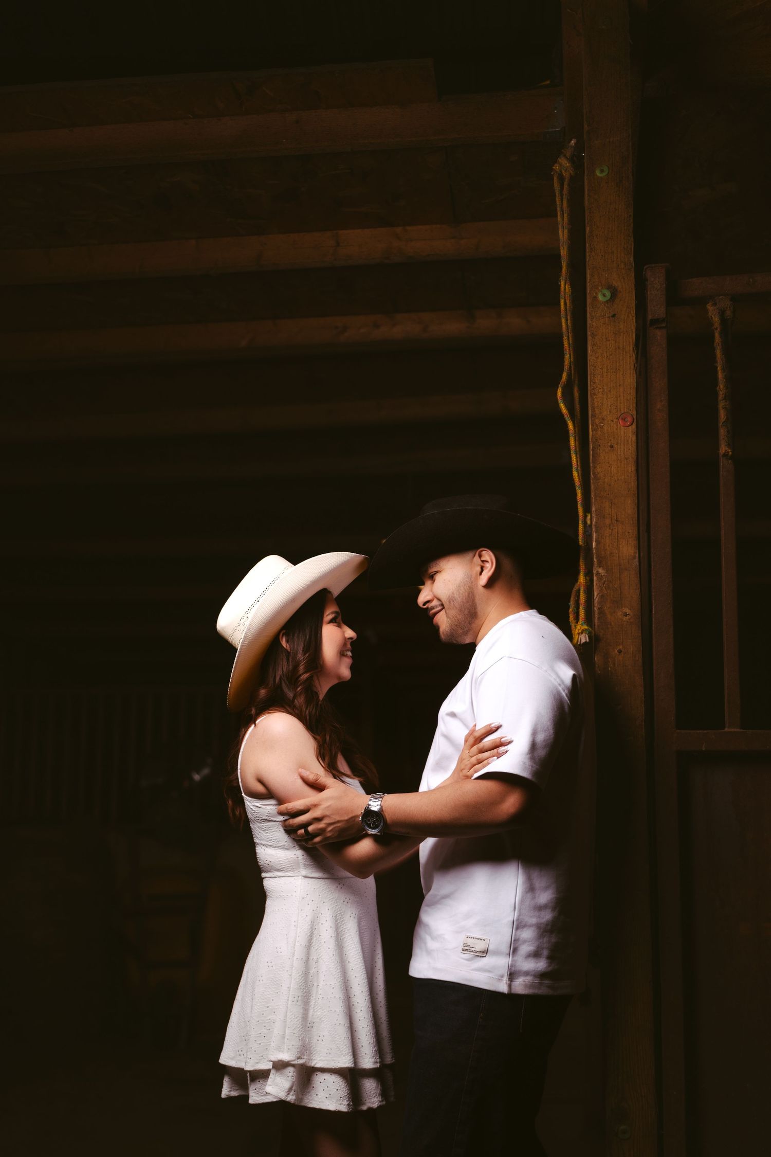 A couple in western hats shares a romantic moment in a rustic barn, smiling at each other while holding hands.