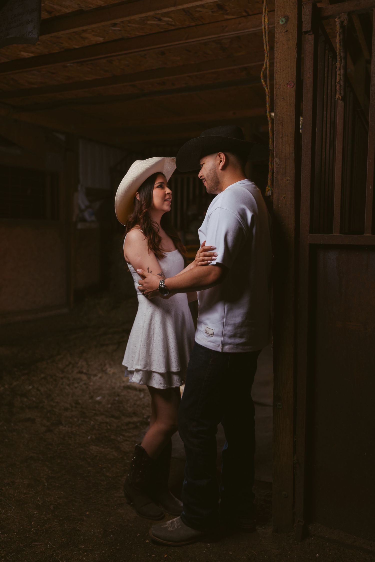 Couple in cowboy hats sharing a romantic moment inside a dimly lit rustic barn.