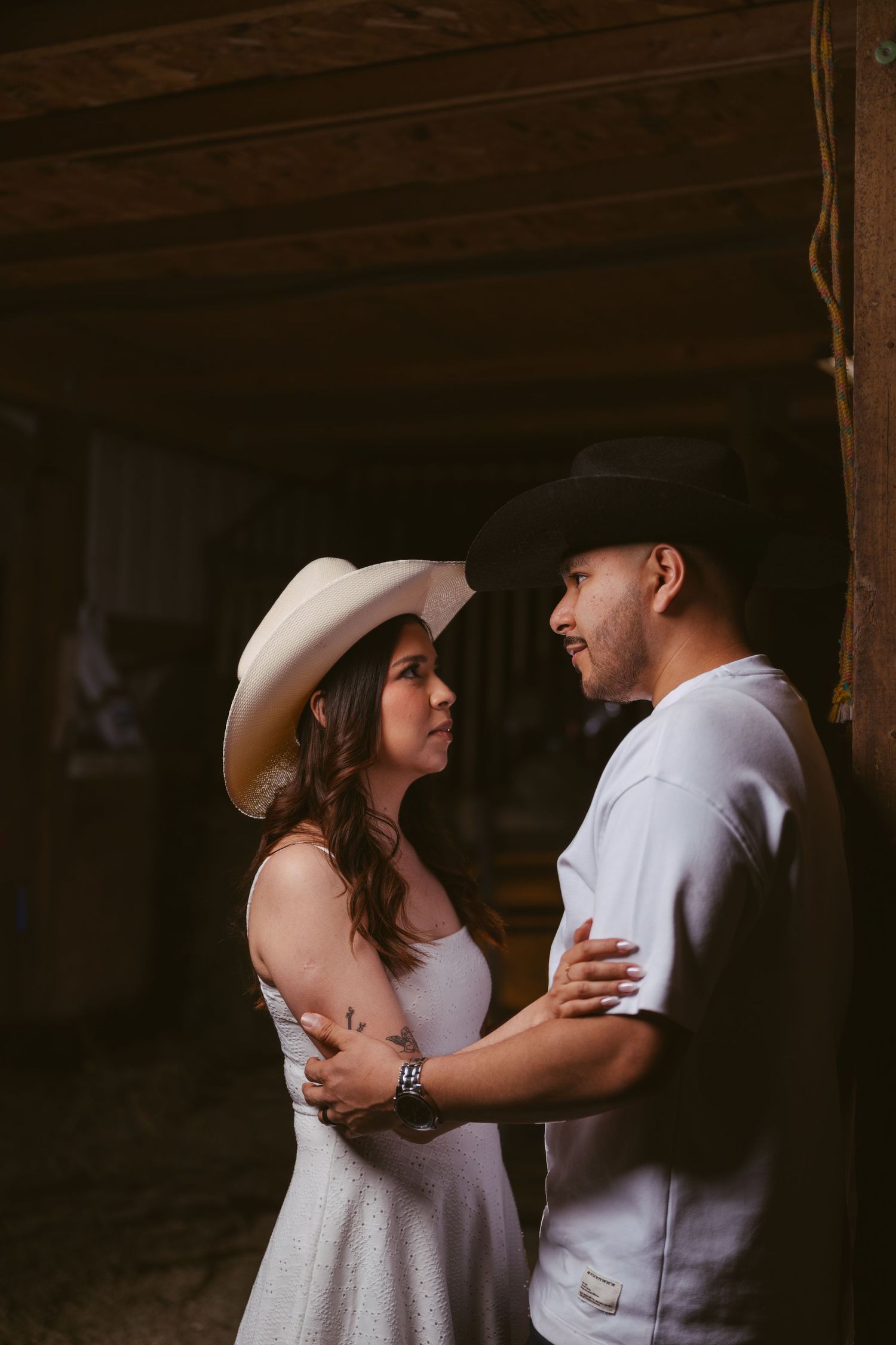 Couple in cowboy hats share a romantic moment in a rustic barn setting.