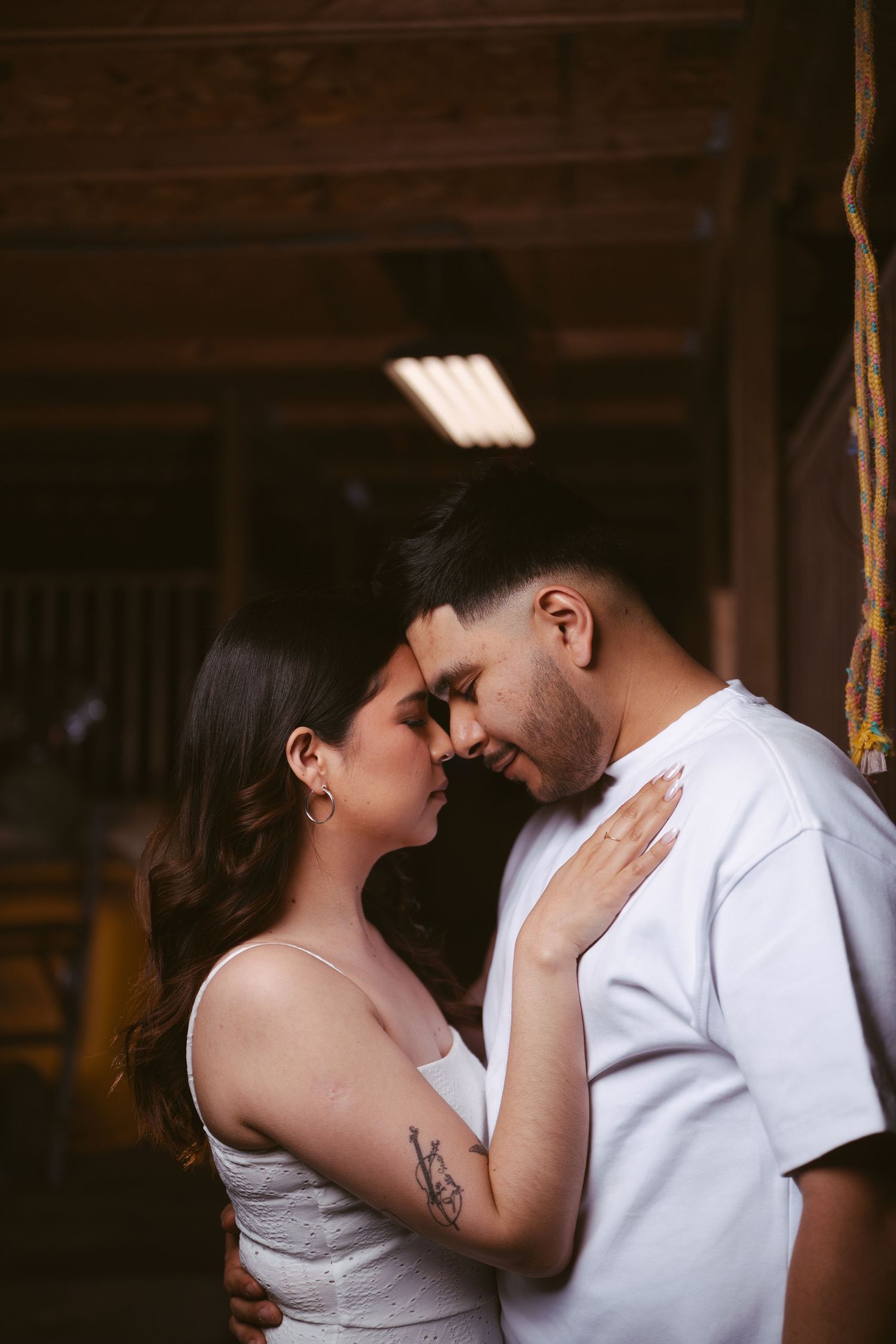 Couple sharing an intimate forehead-to-forehead moment in a rustic barn, woman in white dress with tattoo.