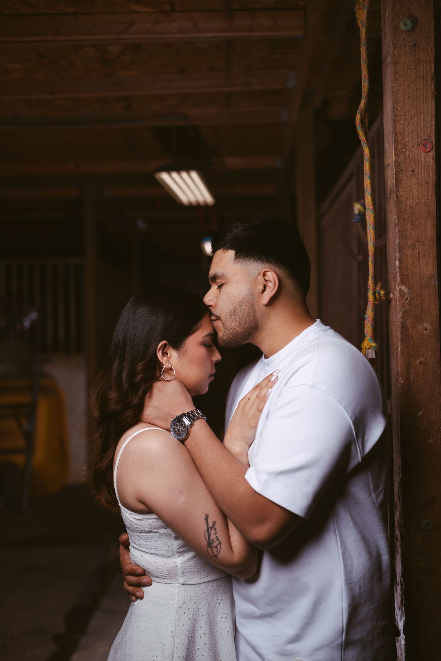 Couple sharing an intimate moment leaning against a wooden barn door, woman in white dress with tattoo.