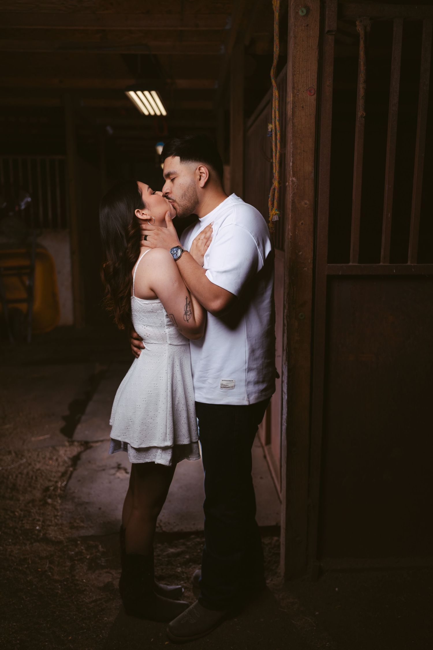 Romantic couple kissing in a dimly lit barn hallway, man in white t-shirt and woman in white dress.