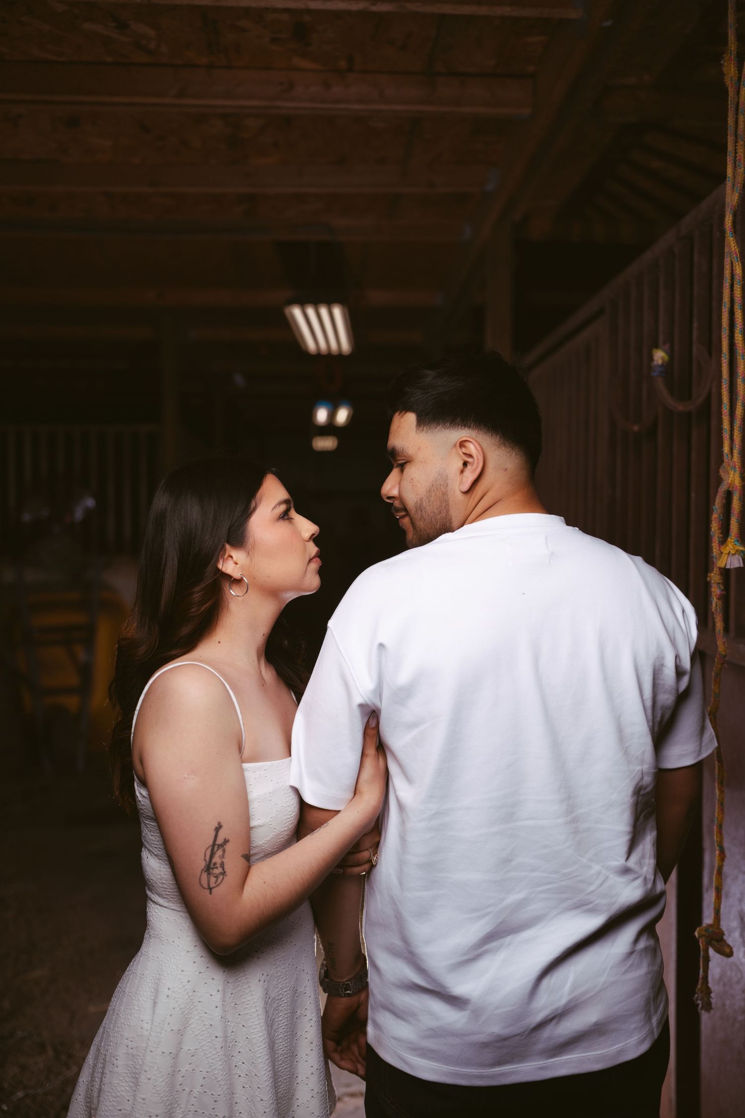 Couple sharing an intimate moment inside a rustic barn, woman in white dress with tattoo holding mans arm.