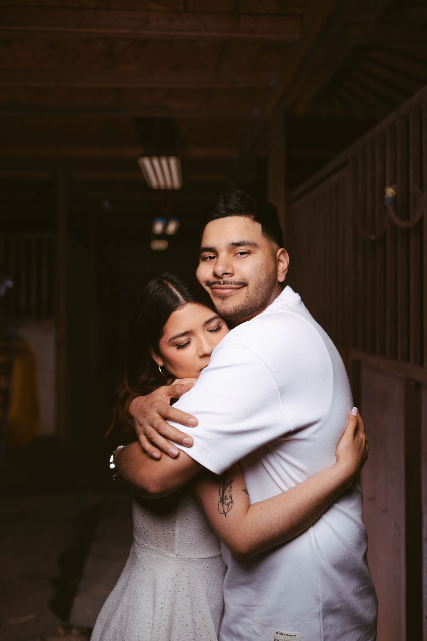 Couple embracing warmly in a barn hallway, woman in white dress hugging man in white shirt.
