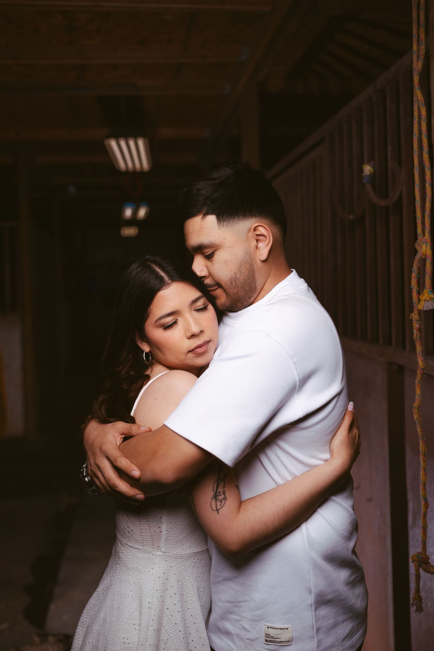 Man tenderly embracing woman from behind in a dimly lit barn, both wearing white outfits.