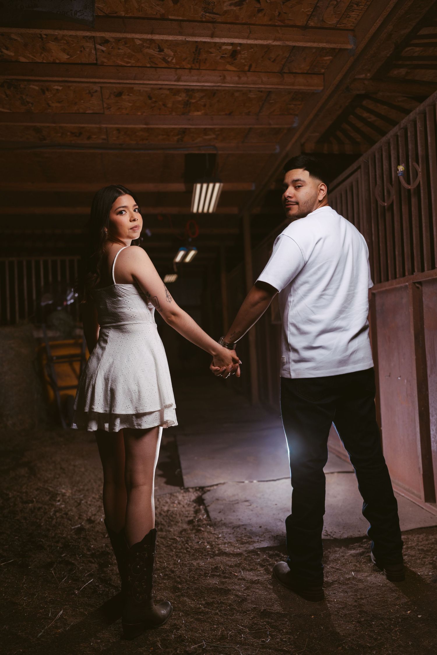 Couple holding hands in a rustic barn, woman in white dress and boots looking back over shoulder.