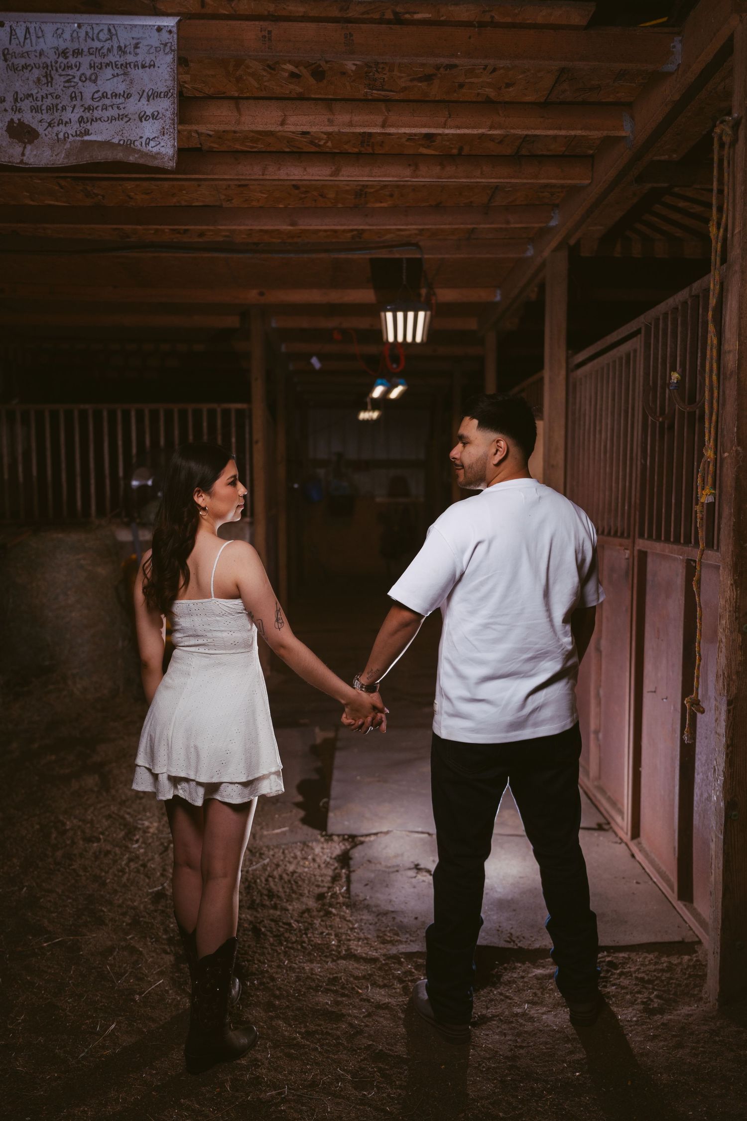 A couple holds hands in a rustic barn aisle, the woman in a white dress and cowboy boots.