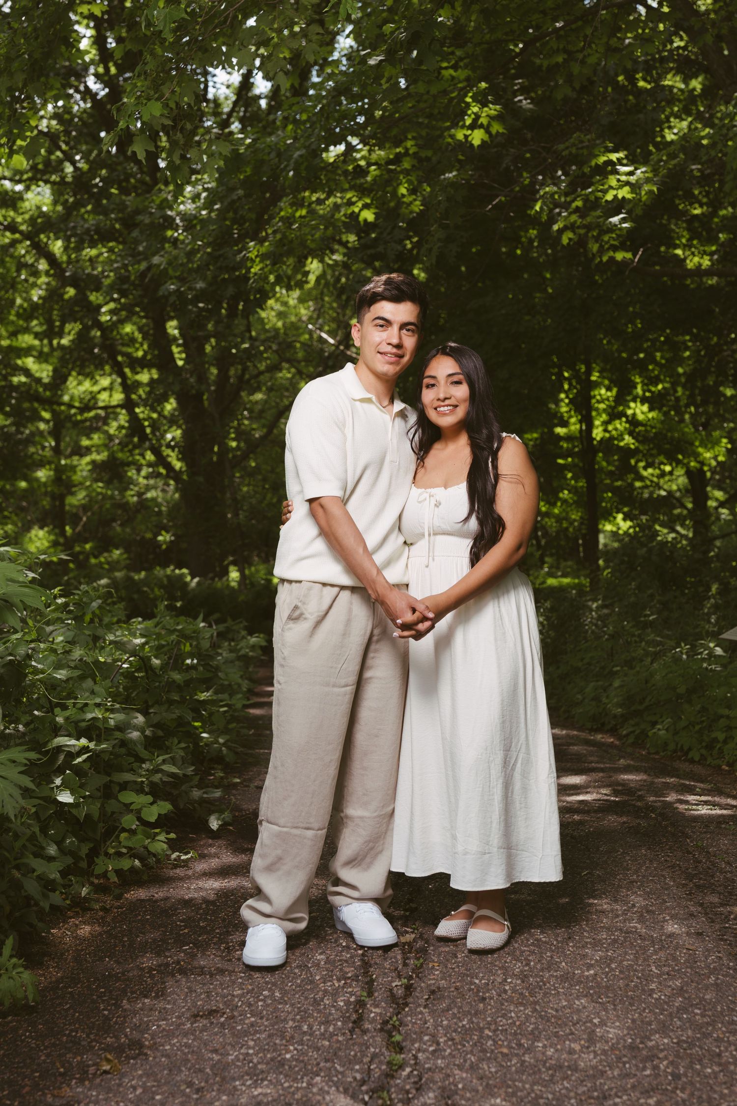 A young couple in matching white outfits holds hands on a forest path surrounded by lush green trees.