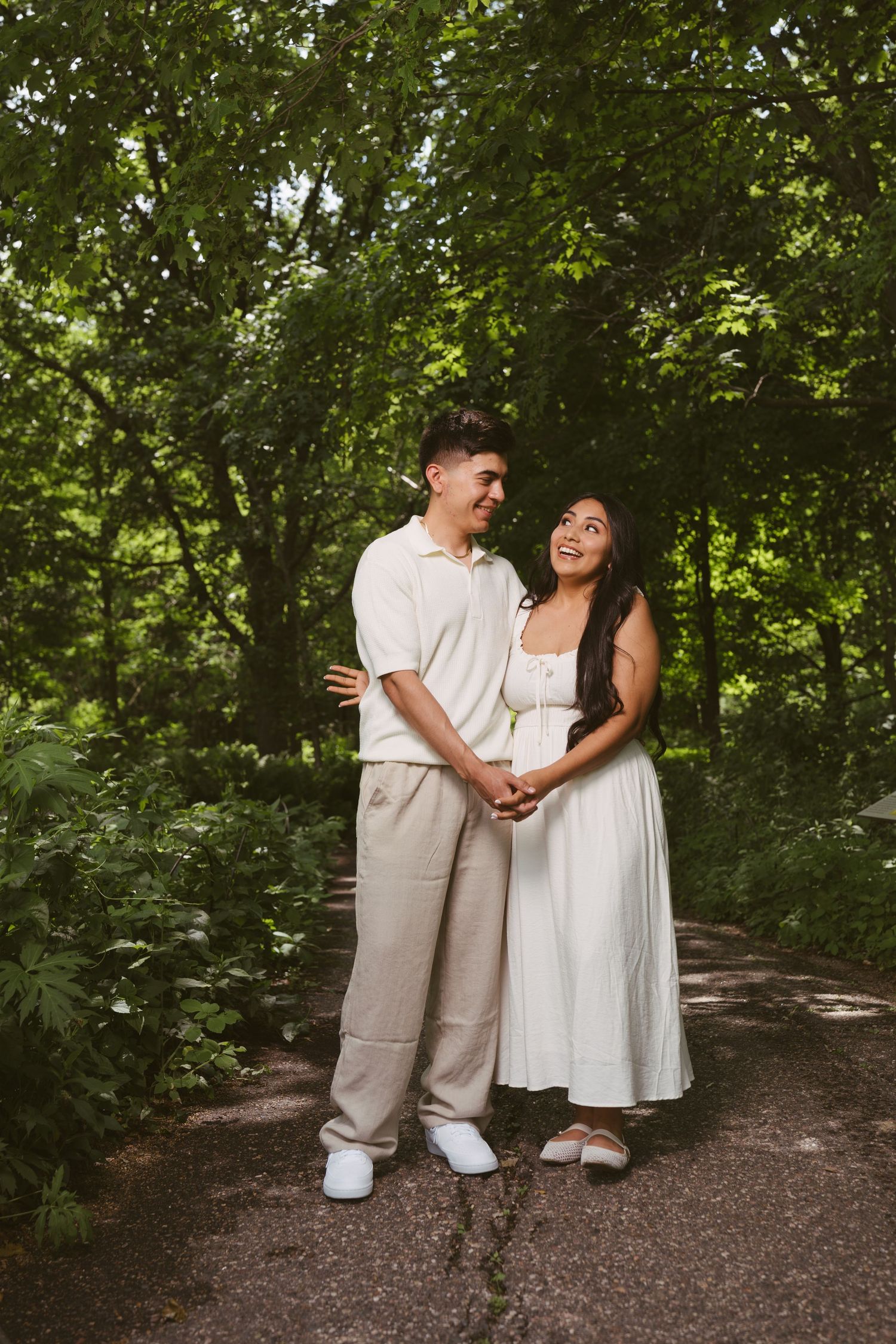 A happy couple holds hands and smiles at each other on a lush green tree-lined path in coordinating neutral outfits.