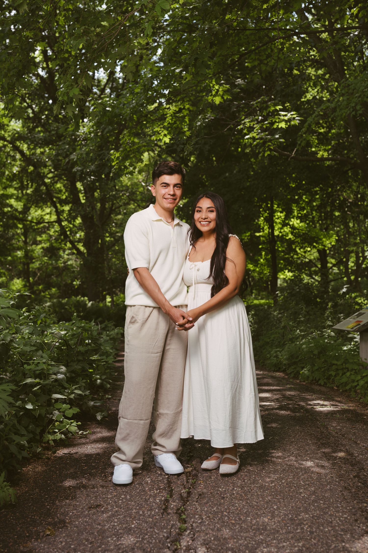 Young couple in neutral outfits holding hands on a tree-lined path in a lush green park.