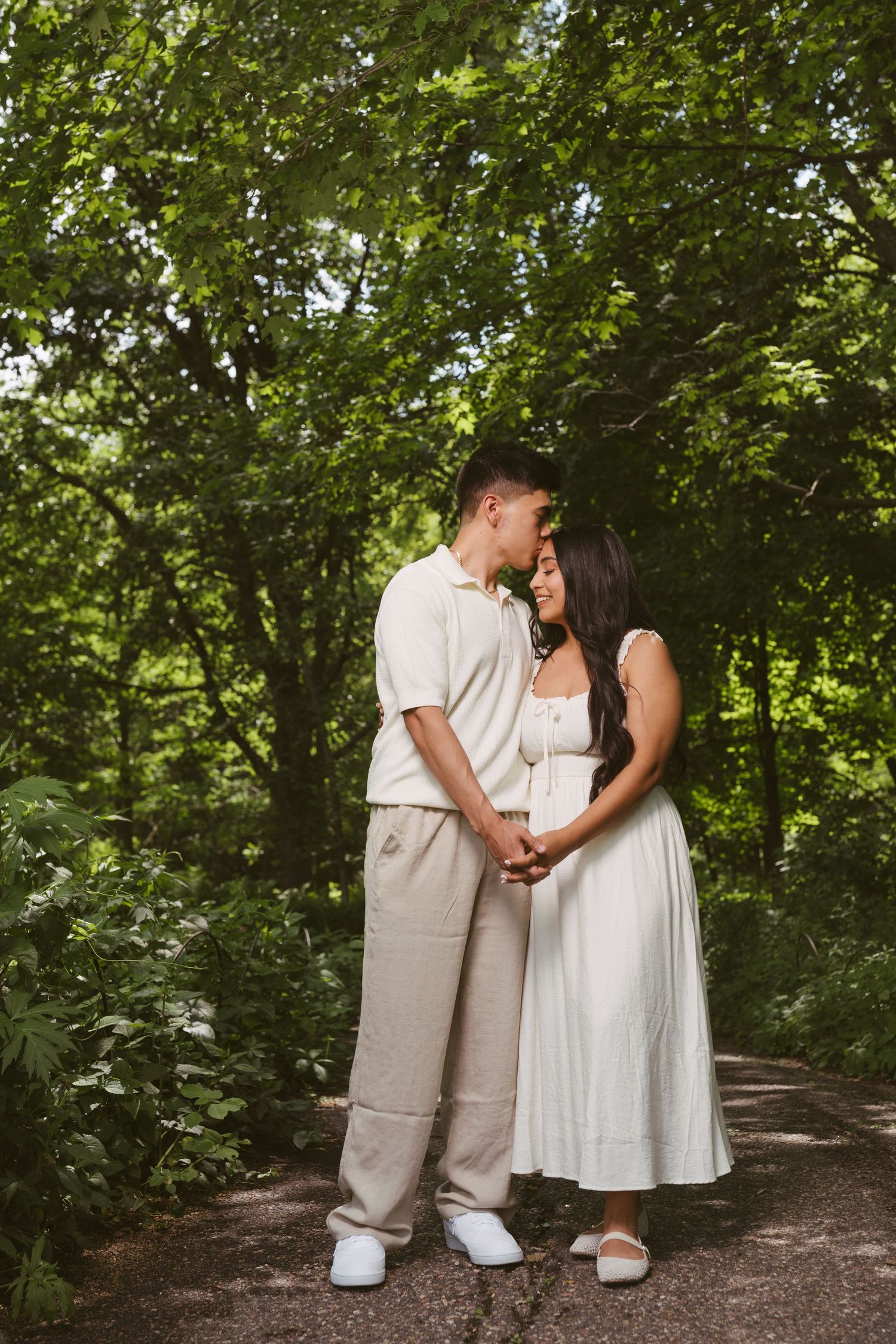 Couple in matching cream outfits holding hands and sharing a kiss on a forest path surrounded by lush green trees.