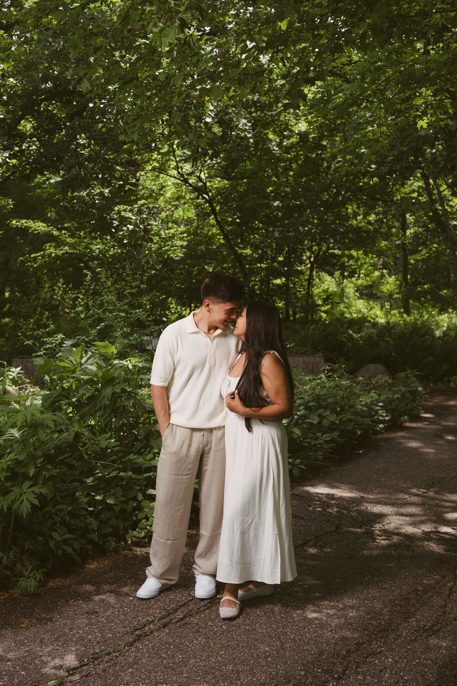 Couple sharing a tender moment on a lush green park path, woman in white dress leaning into man in beige outfit.