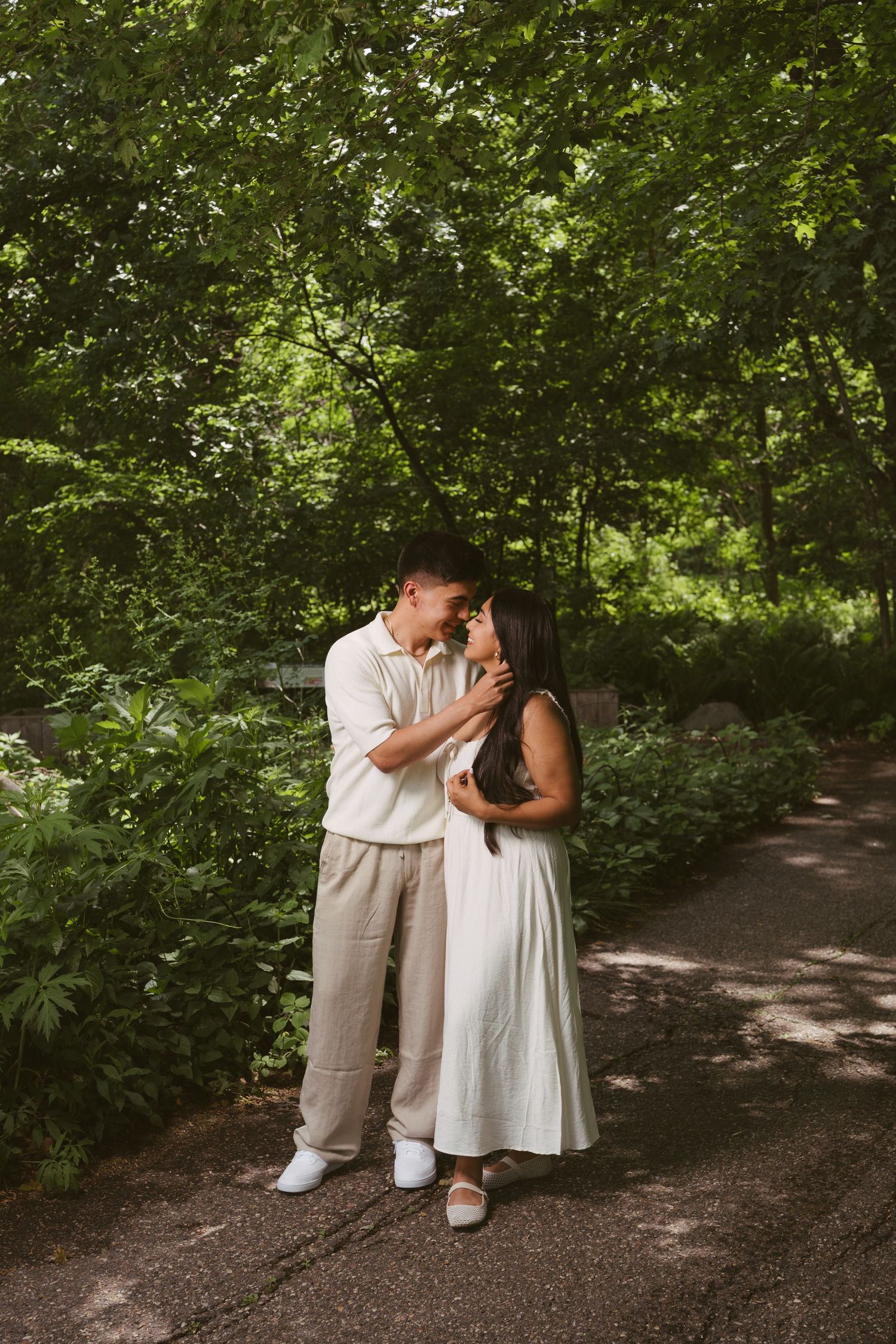 Couple sharing a romantic moment on a tree-lined path, woman in white dress, man in beige outfit.